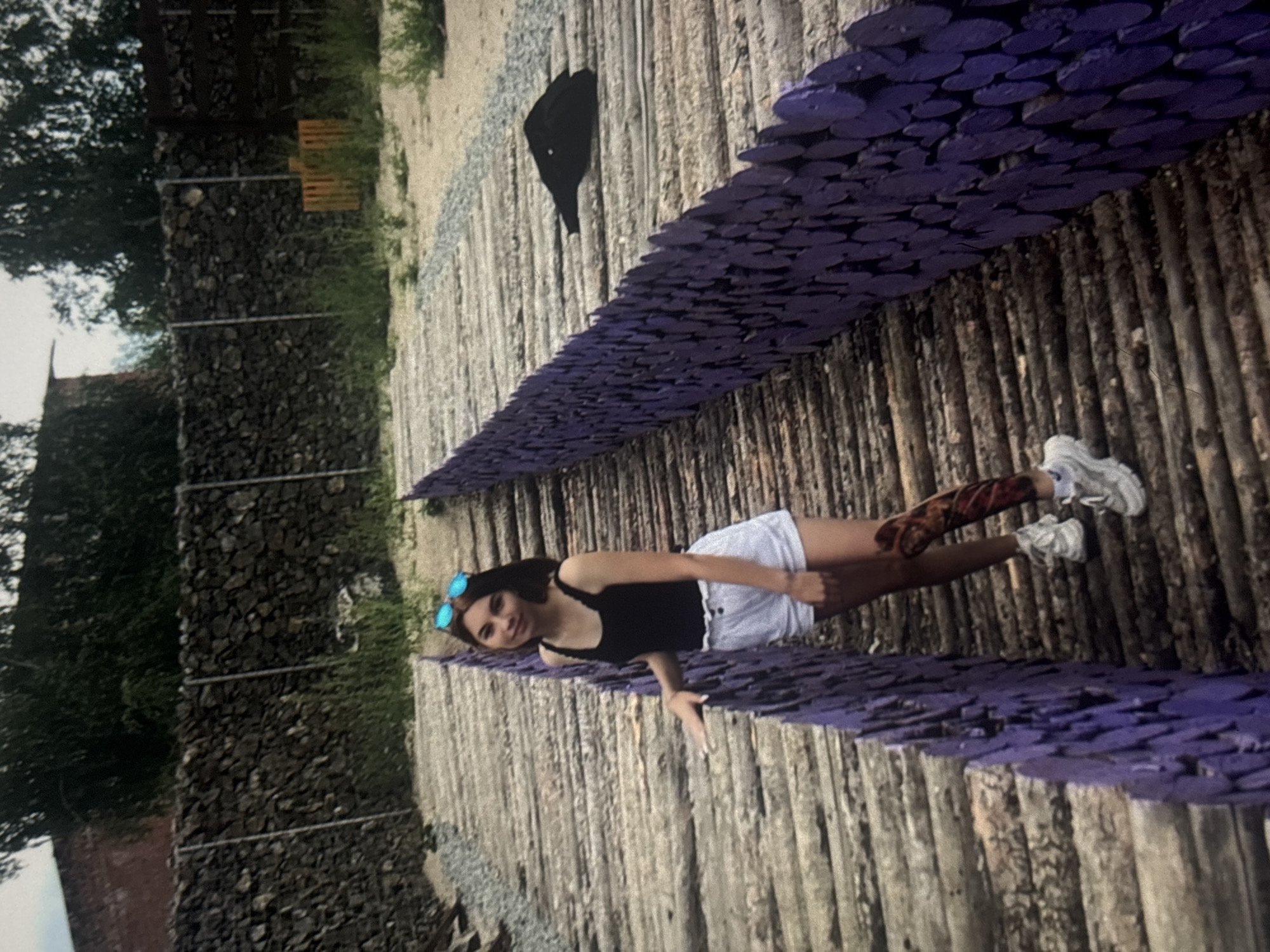 A young woman stands on a wooden walkway with a purple wooden fence on her right. She is wearing a black tank top, white shorts, and white sneakers. She has sunglasses on her head and a tattoo on her right leg. The walkway is made of logs and is in an outdoor setting with a stone wall in the background. The location is likely a park or recreational area in the city of Omsk, Russia. A black jacket is lying on the walkway to the right of the fence.