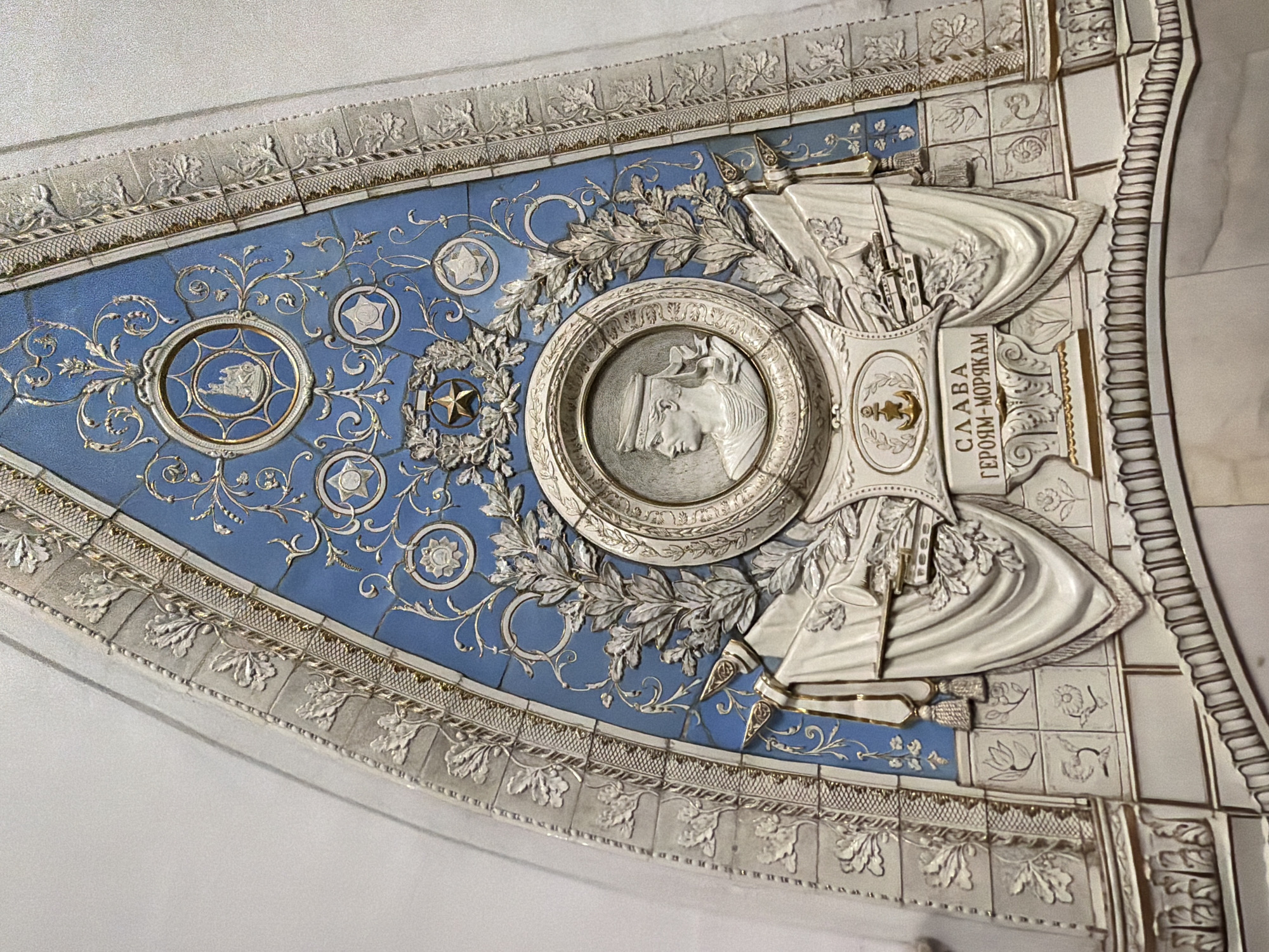 This image shows a triangular-shaped, ornate tile panel. The panel has a blue background with intricate white and gold decorative elements, including stylized floral patterns, circular motifs, and stars. At the center of the panel is a circular medallion containing a profile portrait of a woman in relief. To the right of the medallion is a raised white section with crossed flags, rifles, and the inscription "СЛАВА ГЕРОЯМ-МОРЯКАМ" ("Glory to the Heroic Sailors"). The panel is bordered by a white, decorative frame with leaf patterns and geometric accents.
This tile panel is located in the Komsomolskaya station of the Moscow Metro in Russia.