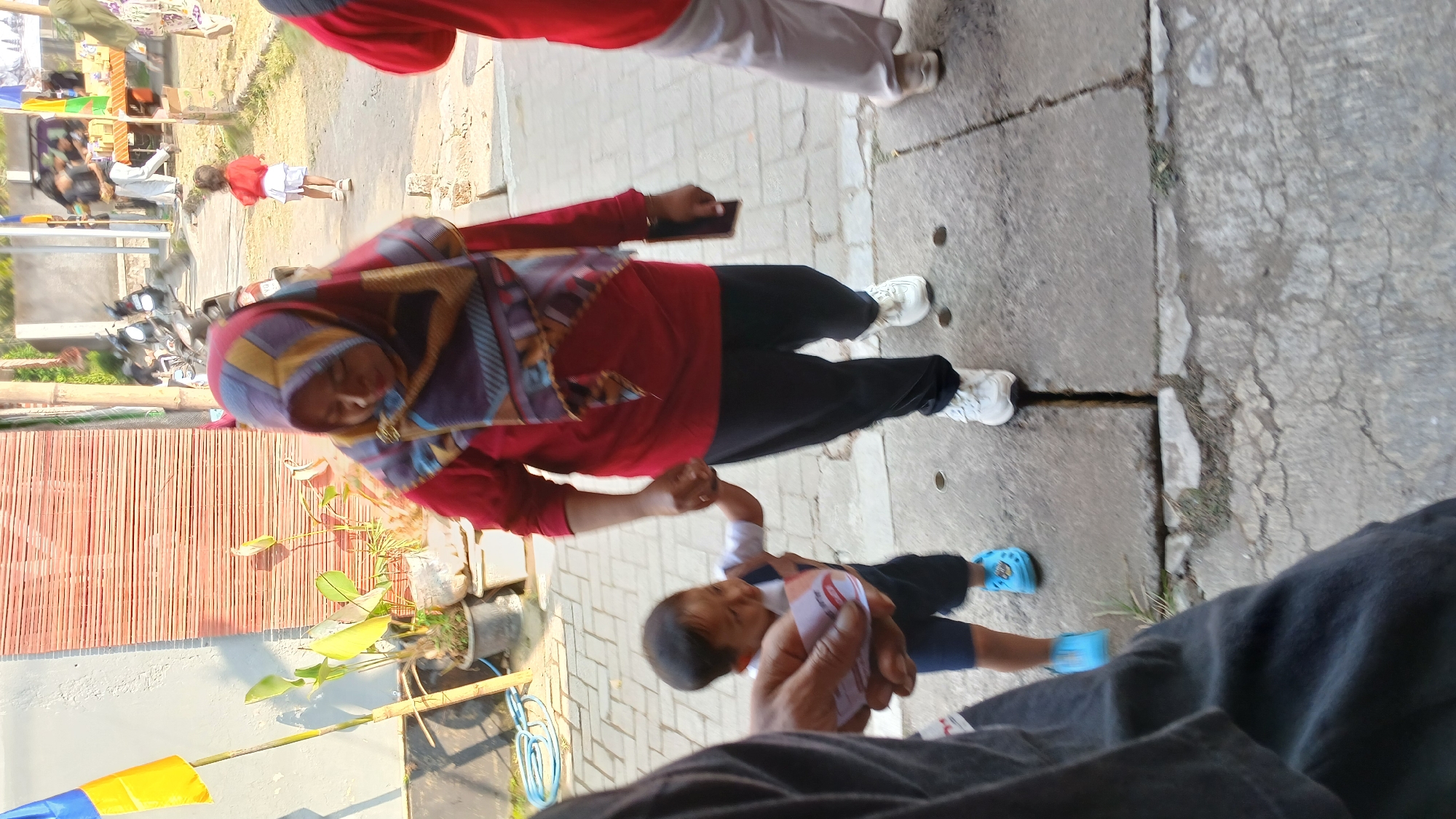 In Mendut, Indonesia, a woman wearing a red shirt and a patterned hijab walks on a paved walkway, holding the hand of a young child. The child, dressed in a white and blue outfit and blue Crocs, is looking down at a card held in their hand. The background shows a building with a textured wall and horizontal wooden slats, plants in pots, and some distant figures and parked motorcycles. The lighting suggests it is daytime with sunlight casting shadows.