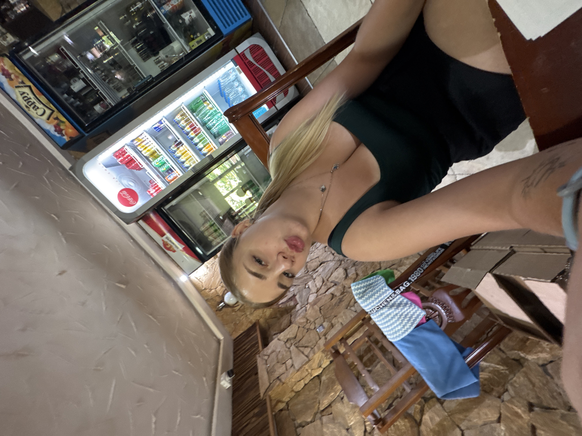A young woman with blonde hair pulled back is taking a selfie in what appears to be a restaurant or cafe. She is wearing a dark green tank top and a black skirt. She is seated at a wooden table, with a stone wall behind her. In the background, there are two glass-fronted refrigerators filled with drinks. One refrigerator has a "Cappy" logo at the top and the other has a "Coca-Cola" logo. The woman's face is tilted, and she is puckering her lips. A wooden chair is to her left, with a folded blue cloth and a striped towel draped over it. The overall lighting is warm, suggesting indoor illumination. The location is identified as Sredets, Bulgaria.
