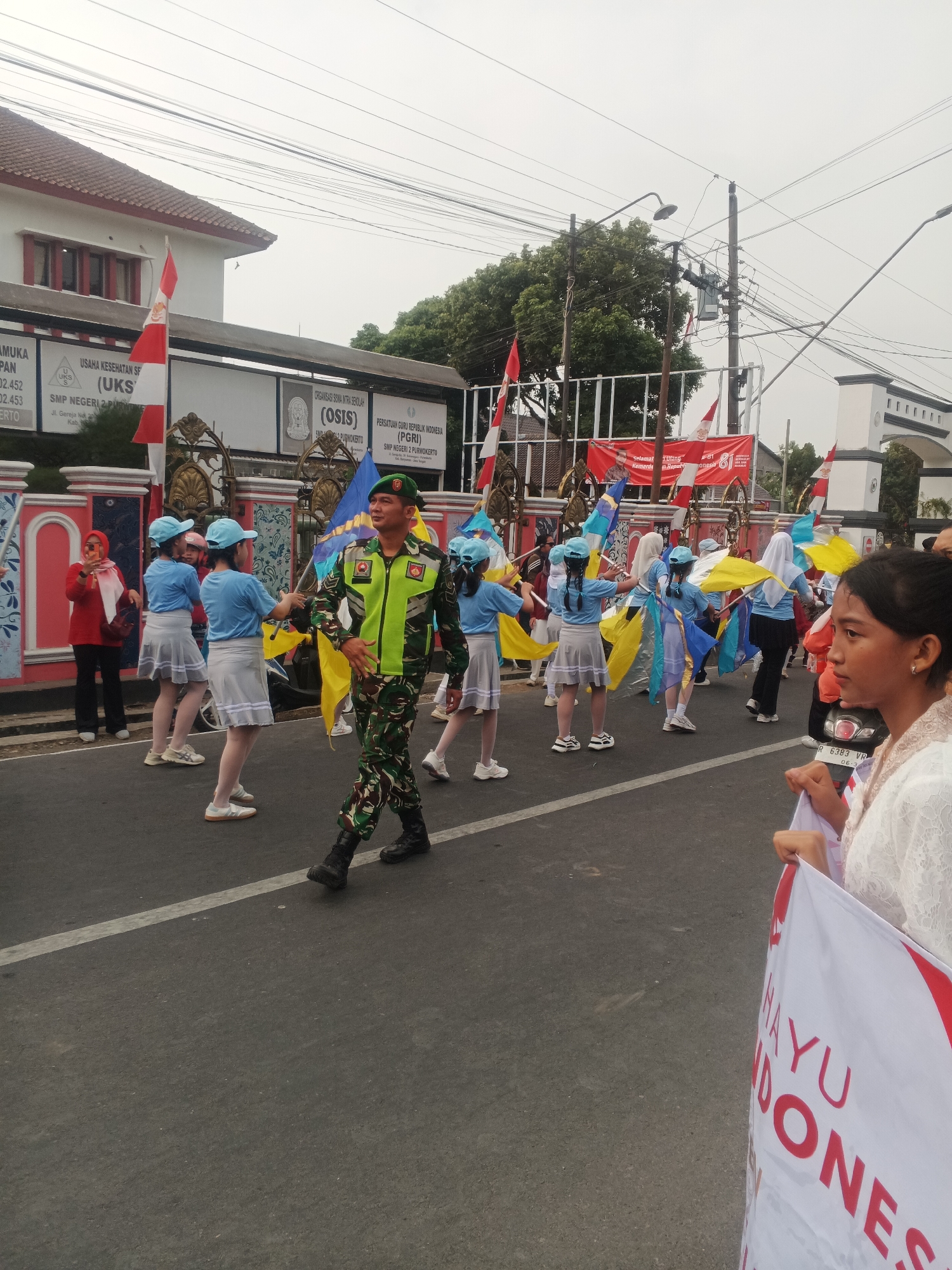 The image depicts a street scene in Sokanegara, Indonesia, possibly during a parade or event. A group of young girls in blue shirts and grey skirts are performing with colorful flags. A soldier in camouflage uniform with a green vest is marching in the foreground. In the background, buildings with red roofs and white walls are visible, along with banners and flags, including the Indonesian flag. The street is paved, and there are power lines overhead. A woman in a red headscarf is standing to the left, holding a phone. A young woman in a white lace garment is in the foreground on the right, holding a banner with red lettering. The sky is overcast.