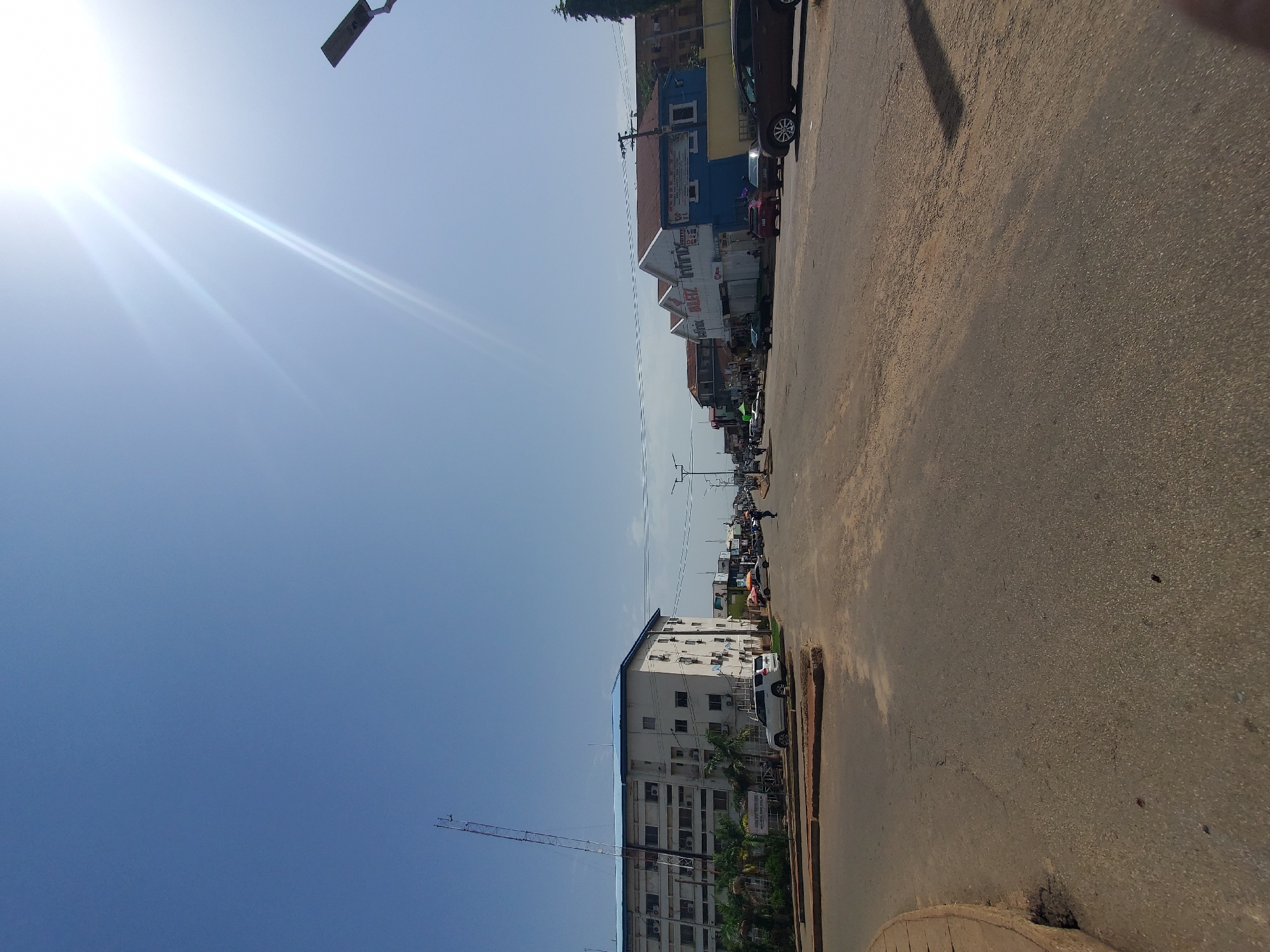 The image depicts a street scene in Jos, Nigeria, on a bright, sunny day. The sky is a clear blue with the sun prominently visible in the upper right quadrant, casting lens flares and bright streaks across the sky. A modern street light is mounted on a pole to the right.
In the foreground, a wide paved road dominates the lower half of the image, showing signs of wear and tear with some cracks and patches. A curved concrete curb is visible on the left side.
On the left side of the road, a large, multi-story building with a blue roof stands. It appears to be an office building or a commercial establishment, with numerous windows and air conditioning units. A tall antenna or communication tower is situated next to this building. Several palm trees are planted in front of the building. A white SUV is parked near the building.
Further down the street, on both sides of the road, are various commercial buildings. Some of these buildings have awnings and signage, indicating shops or businesses. On the right side, there are several cars parked and driving along the road, including a dark-colored SUV, a dark sedan, and a lighter-colored sedan. People can be seen walking on the sidewalks and crossing the street. The overall atmosphere suggests a busy urban environment. The buildings and the style of the street suggest a commercial district within Jos.