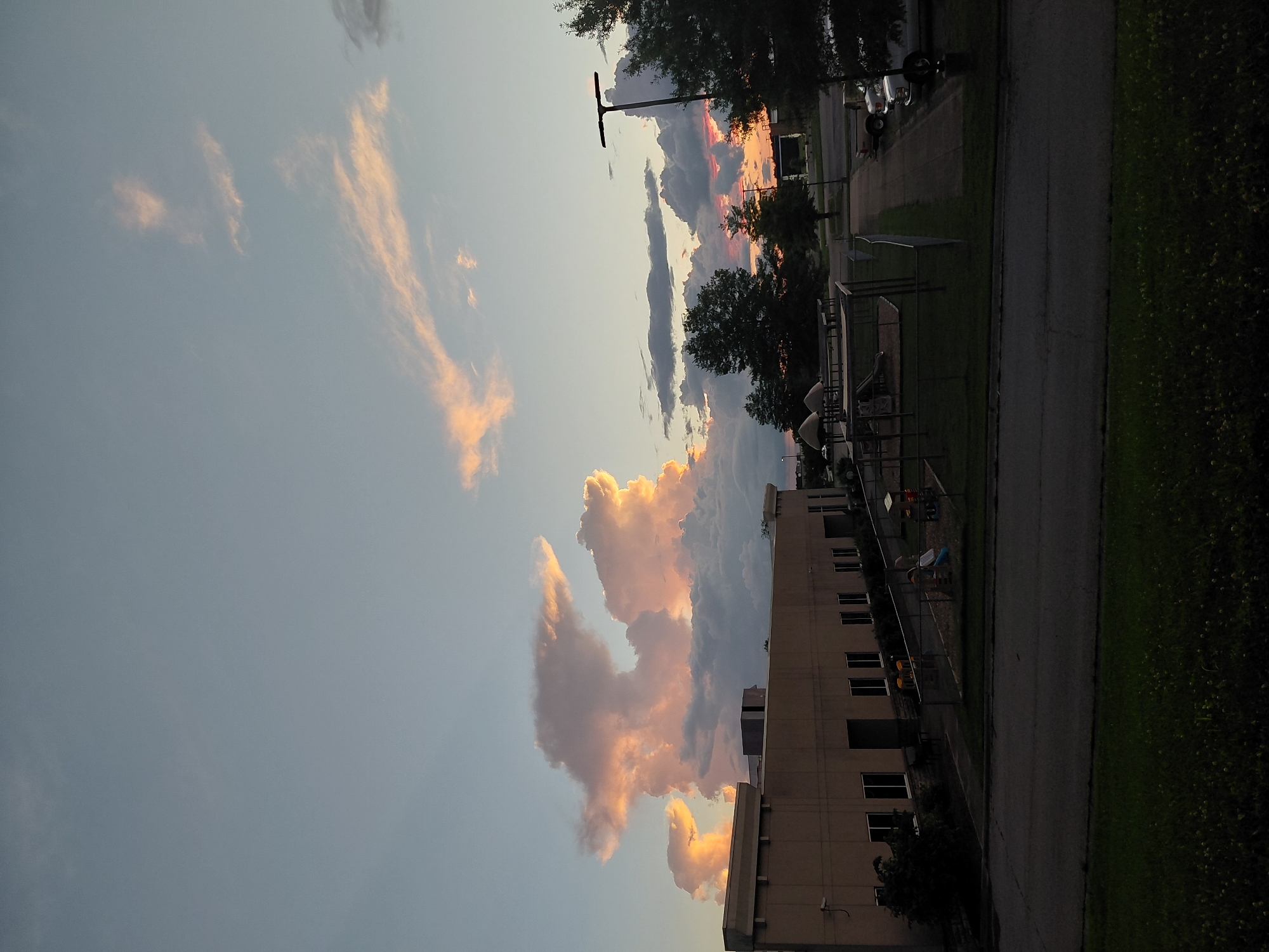 The image depicts an outdoor scene at dusk in Nicholasville, Kentucky, United States of America. The sky is filled with dramatic, cloud formations illuminated by the setting sun, casting pink and orange hues across the sky. In the foreground, a dark asphalt road leads the eye towards a building, likely a school or office, with a beige facade and numerous windows. Adjacent to the building is a fenced-in playground area with swings and slides. Several trees are scattered throughout the scene, with one particularly large, dark tree dominating the right side of the image. A lamppost stands tall near the trees. Parked vehicles, including trucks and cars, are visible on a paved area next to the building. The overall impression is one of a quiet, end-of-day atmosphere, with the natural beauty of the sunset contrasting with the man-made structures.