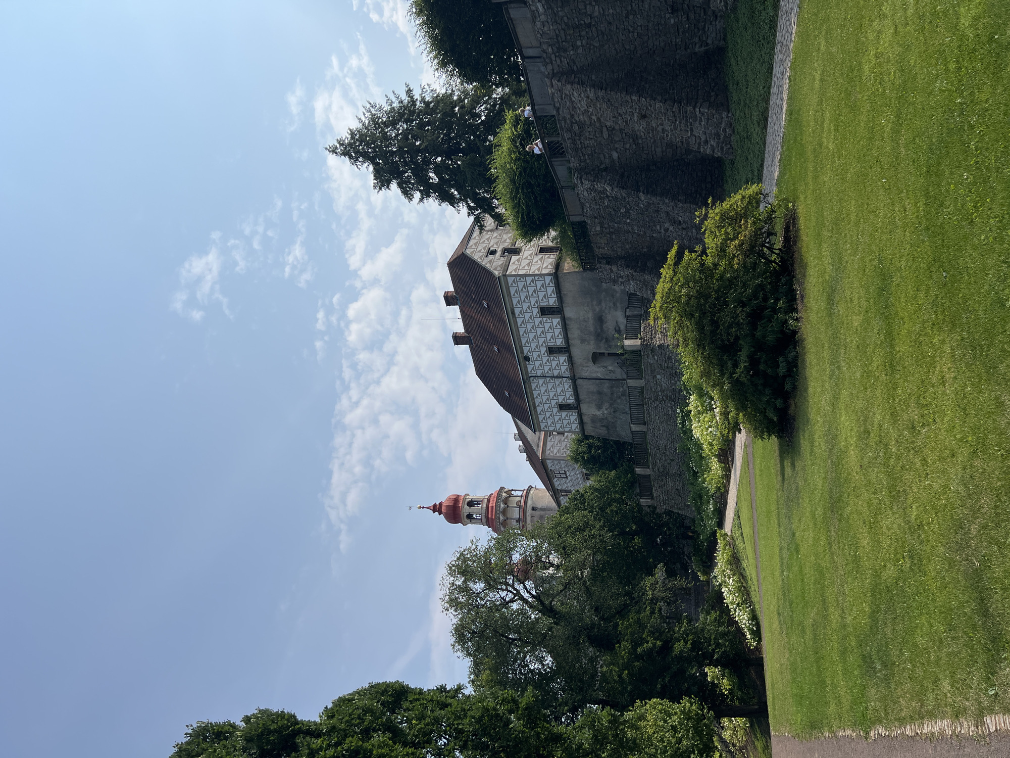 The image depicts a scenic view of Náchod Castle in Náchod, Czechia, on a clear, partly cloudy day. In the foreground, a lush green lawn slopes gently downwards, bordered by a paved pathway on the left. To the right, a large, dense bush sits in the middle of the lawn, casting a shadow. Behind the lawn, a stone wall with a wooden railing extends across the frame. Two people are visible standing on the wall, looking out at the scenery.
Dominating the midground and background is the castle complex. A prominent tower with a distinctive red, multi-tiered roof rises above other buildings. The castle walls feature a repeating geometric pattern in grey and white. Mature trees, including a tall conifer on the right, surround the castle, with some branches obscuring parts of the architecture. White flowers are visible in beds along the pathway leading up to the castle. The sky is a vibrant blue, with scattered white clouds.