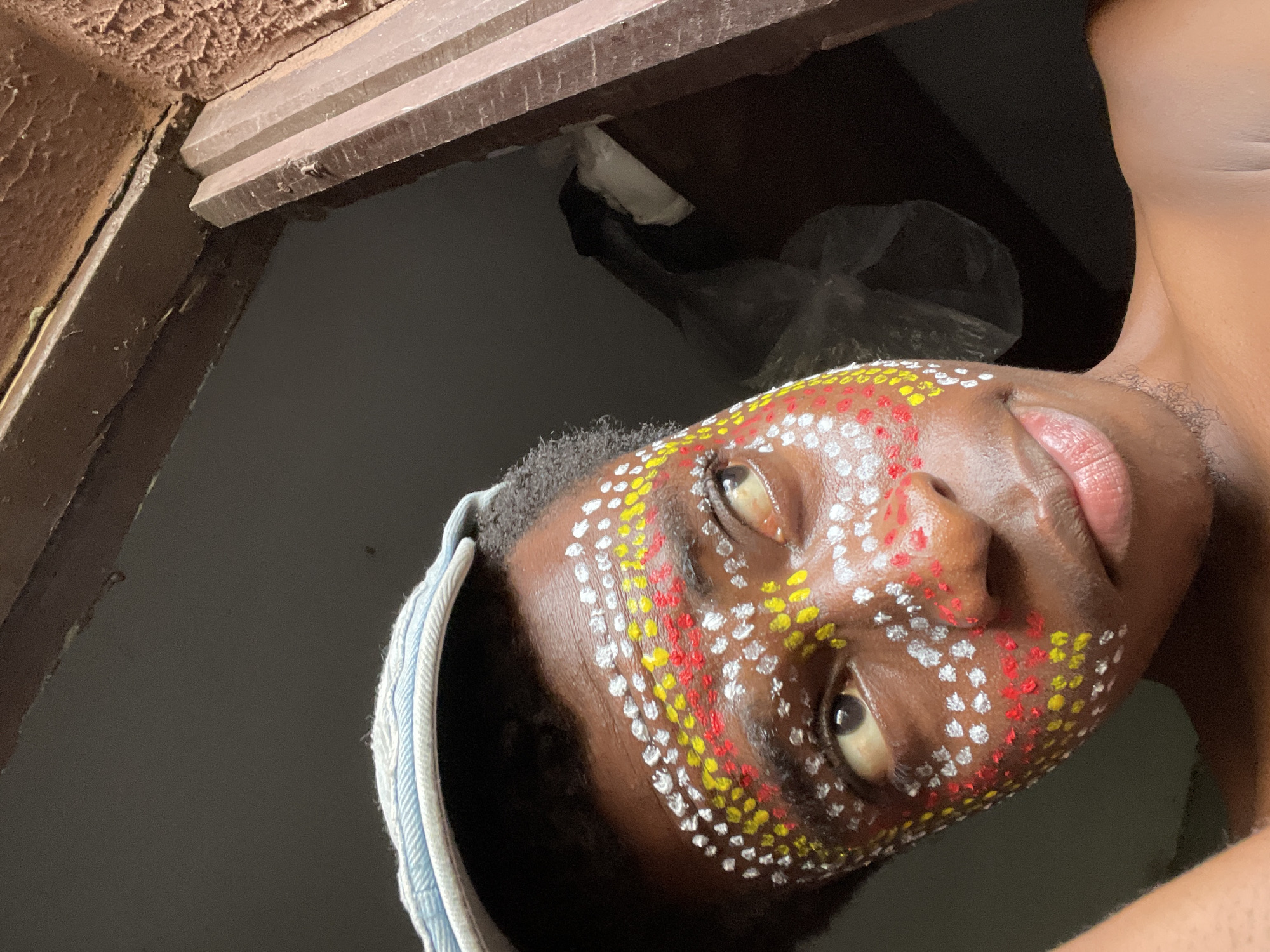 A close-up, tilted shot depicts a dark-skinned individual with white, yellow, and red paint dots on their face, arranged in lines. They wear a light blue headband. The person's left eye is visible, looking directly at the camera. The background is dark, revealing a portion of a door frame and ceiling with textured paint. Underneath the door frame, there are some items. The location is identified as Nkpor, Nigeria.