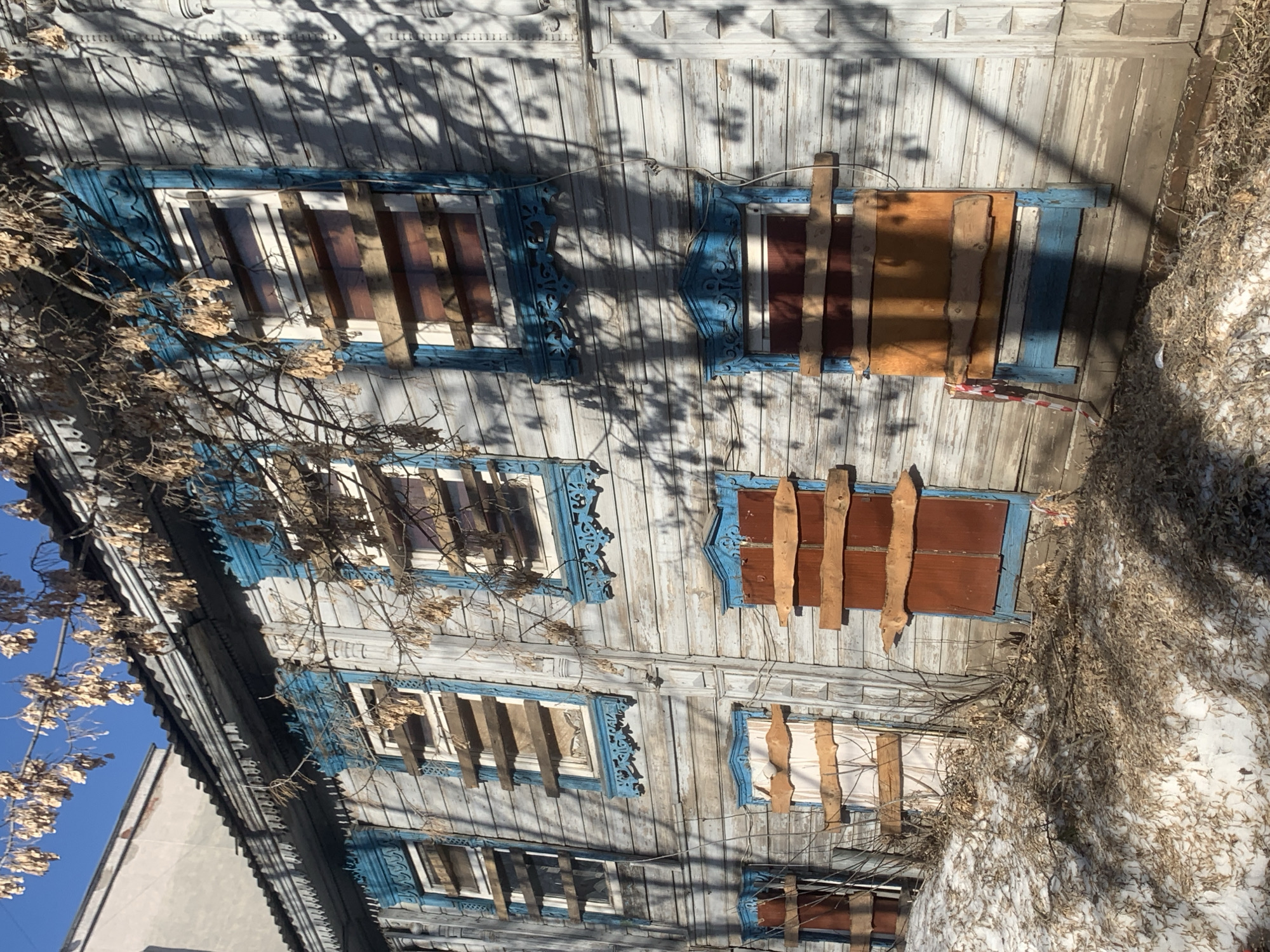 The image depicts the façade of an aged, two-story wooden building, potentially residential, in Izhevsk, Russia. The structure exhibits signs of disrepair, with faded light blue paint on the wooden siding and ornate window frames. All the windows are boarded up with vertically placed wooden planks of varying colors. Snow and dry foliage are present on the ground and around the building. There are bare tree branches partially obscuring the view of the building and a section of a modern grey building can be seen on the left edge.