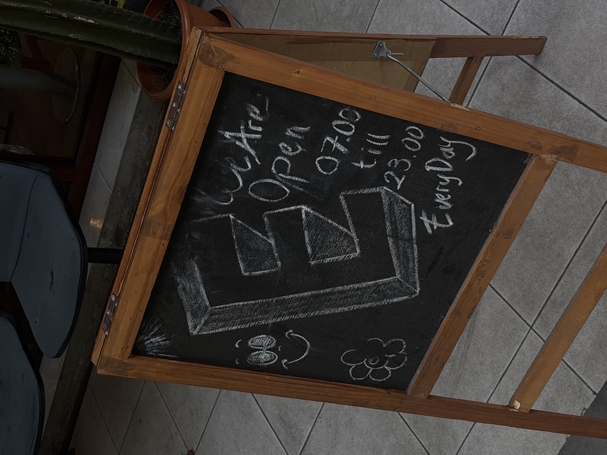 This is an eye-level, medium shot of a wooden A-frame chalkboard sign placed on a tiled sidewalk in front of a building. The chalkboard has chalk drawings and writing on it, including a large, stylized letter "E" with a 3D effect, a smiley face, a flower, and text that reads "We Are Open 07.00 till 23.00 Every Day". The background shows dark blue chairs and a window reflection of a potted cactus. The scene is likely outdoors in a city setting, potentially a commercial establishment in Semarang, Indonesia, given the provided context.