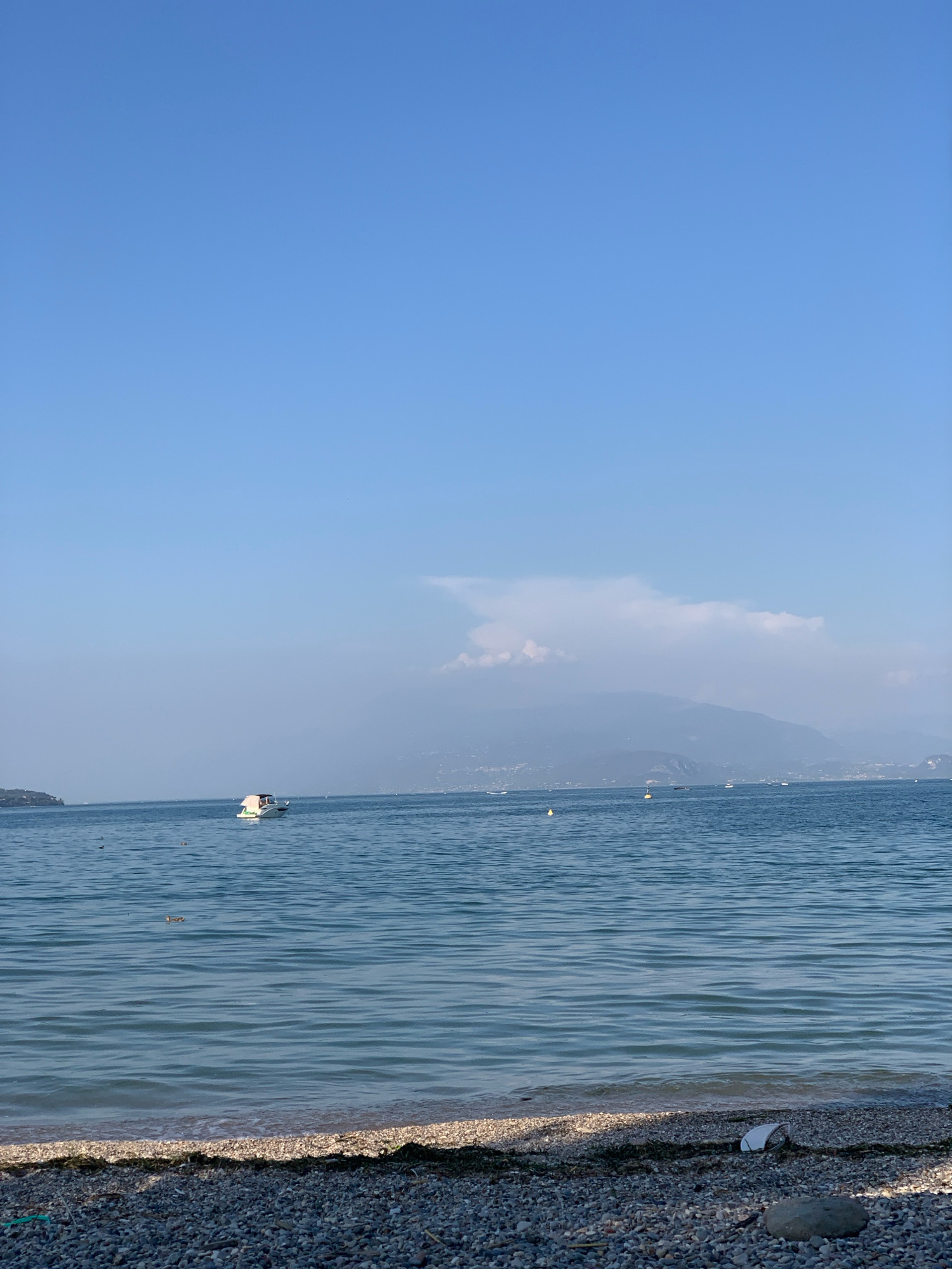 A wide shot captures a serene scene of a clear blue sky over a tranquil body of water, likely Lake Garda, given the proximity to Padenghe sul Garda, Italy. The water's surface is calm, with gentle ripples reflecting the sky. In the distance, a white boat with a beige canopy is visible, appearing to be anchored or moving slowly. Several other smaller buoys and distant boats dot the horizon. Beyond the water, a hazy mountain range is faintly discernible against the sky, with a wispy cloud formation near its peak. The foreground is a pebbled beach, with scattered seaweed and a few rocks, including a distinct grey stone and a white, crumpled object, possibly trash. The overall impression is one of a peaceful, sunny day by the lake.