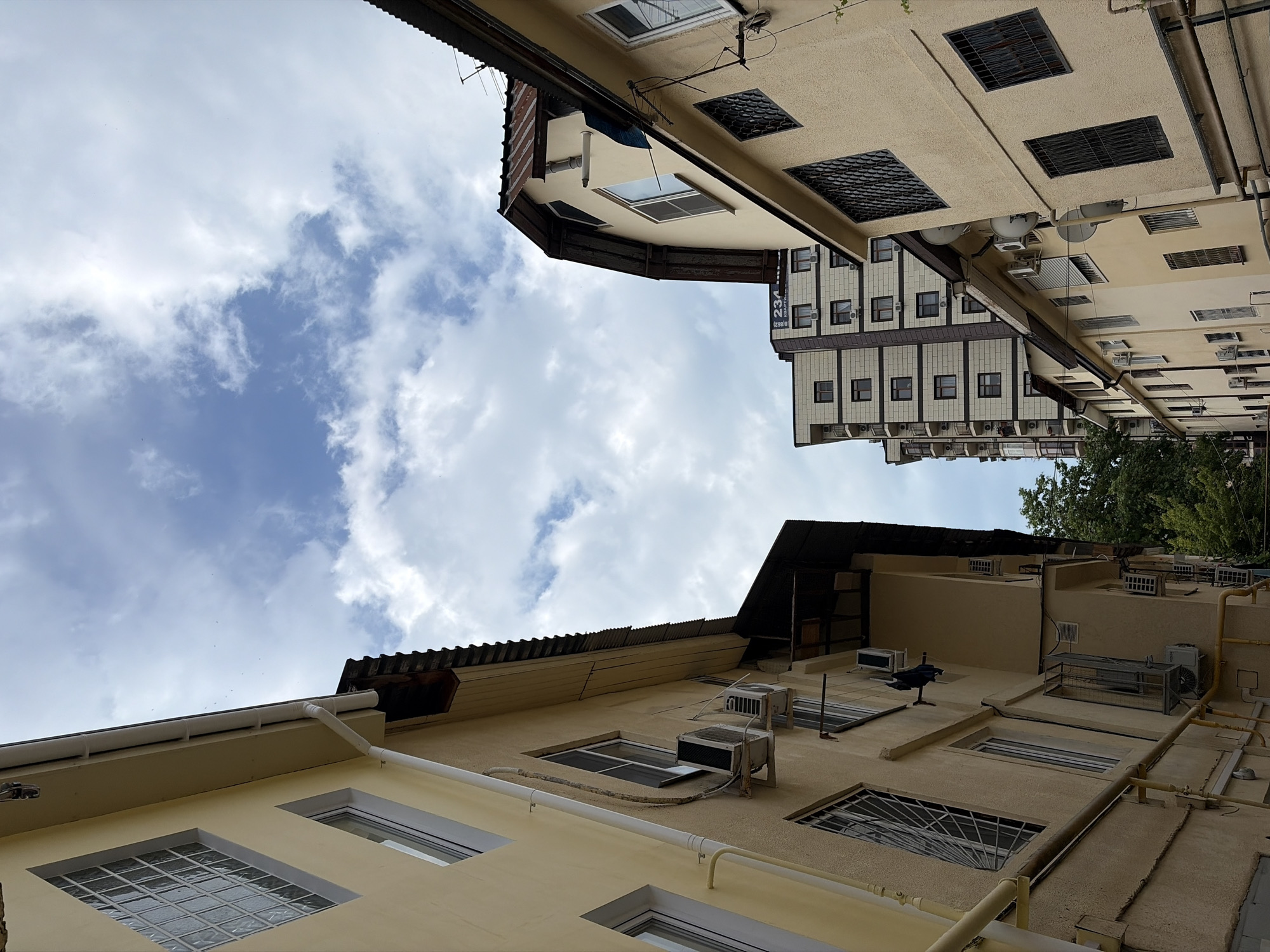 The image shows a view looking up at buildings and the sky in Sochi, Russia. On the left, a pale yellow building with several windows and air conditioning units is visible. To the right of this, a darker, lower building with a sloped, weathered roof is seen. Further to the right and in the background, a taller, modern apartment building with a patterned facade and balconies stands. Green trees are visible behind the lower buildings. The sky is a mix of blue and white clouds. The scene depicts an urban residential environment on a cloudy day.