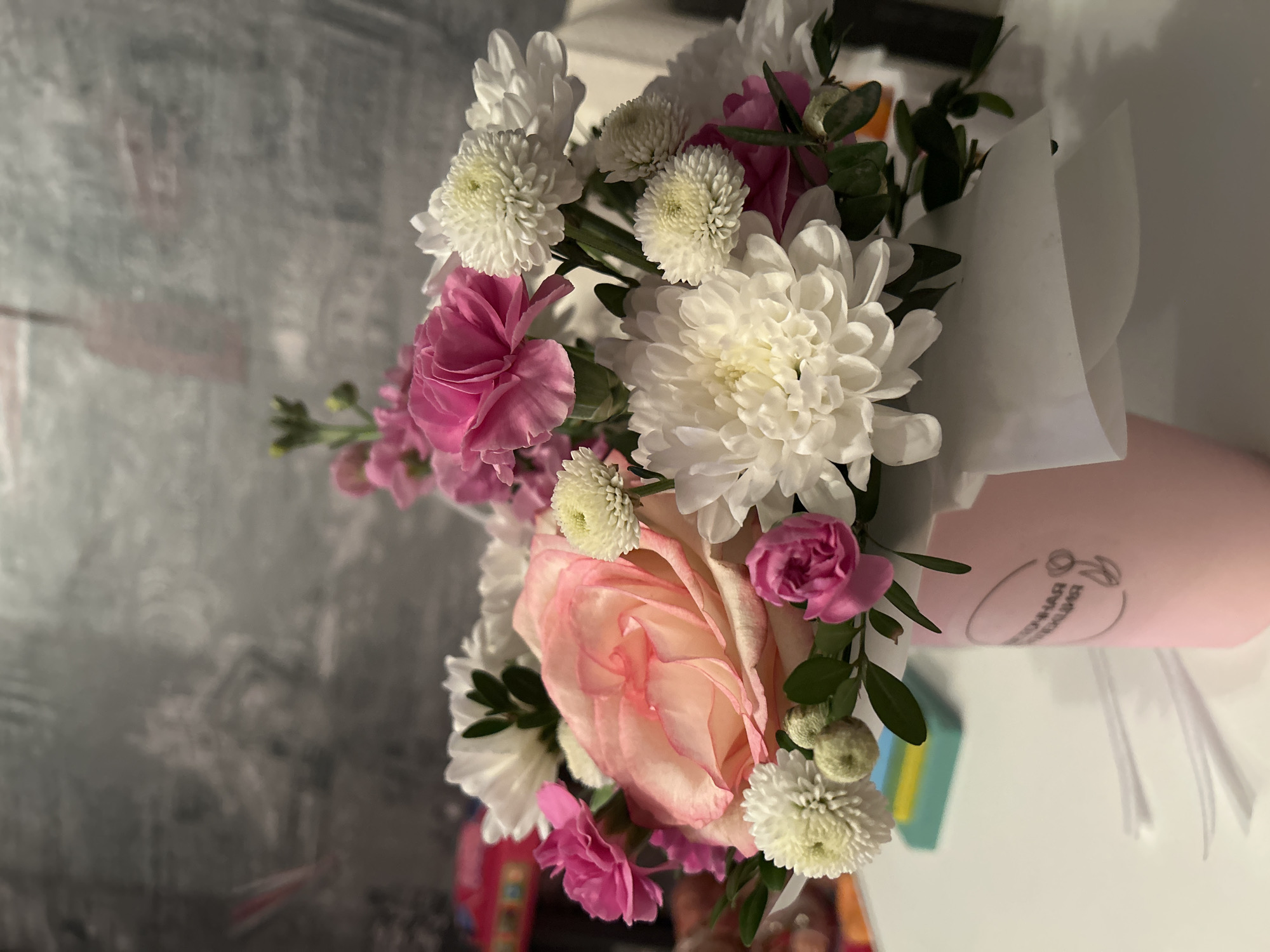 The image features a bouquet of flowers, primarily consisting of white chrysanthemums, pink carnations, and a peach-colored rose, arranged in a pink conical container with white paper wrapping. The location appears to be an indoor setting, possibly in Chaikovsky, Russia, based on the Cyrillic text on the container, which reads "Ручная коллекция." The background is a blurred gray wall. There are no people visible in the image. The primary subject is the bouquet itself, displayed on a white surface.