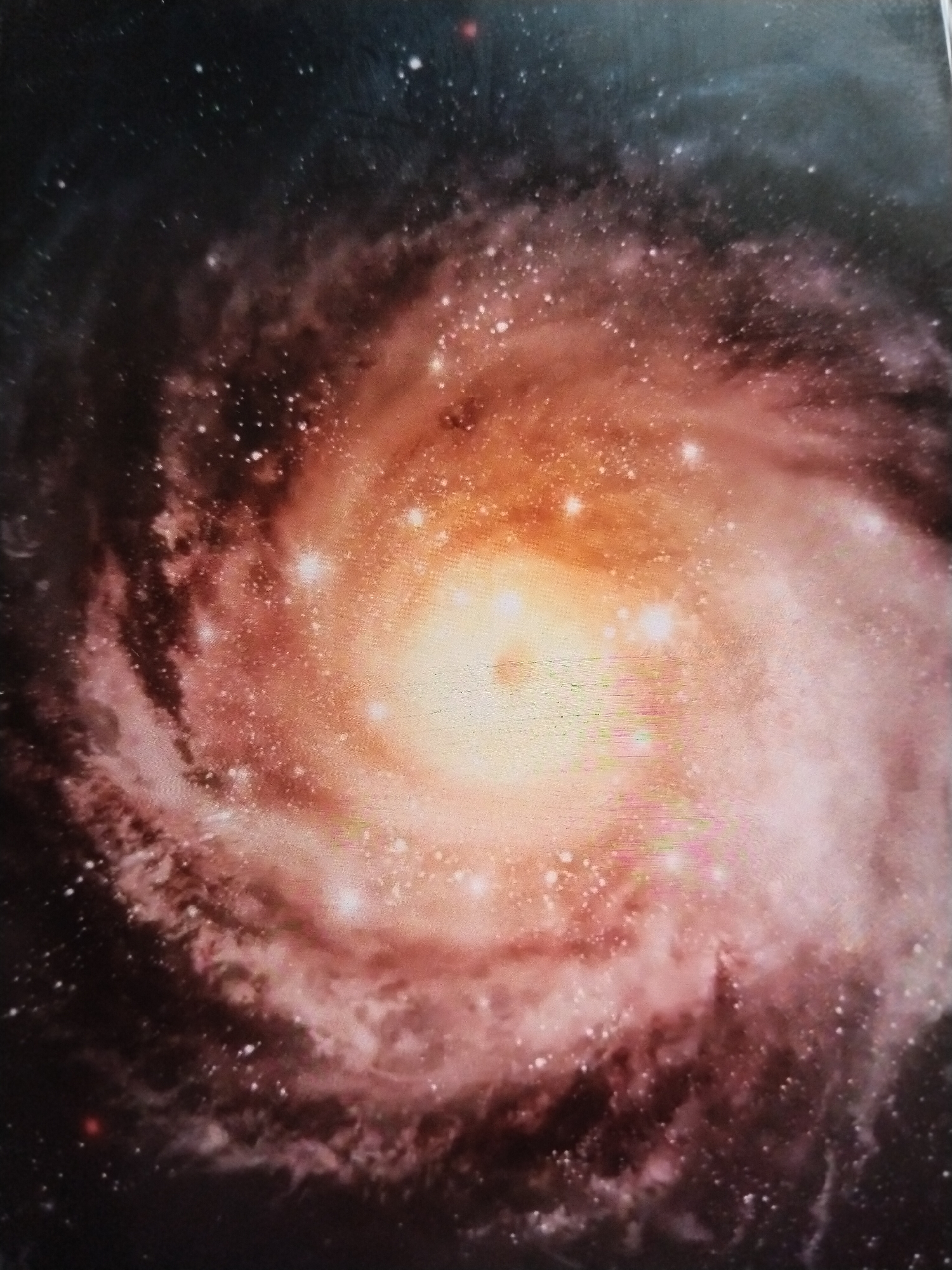 This is an image of a spiral galaxy with a bright, luminous core. The galaxy is depicted with swirling arms of dust and stars, rendered in shades of pink, orange, and yellow, set against a dark, star-filled background. There are no people, objects, actions, interactions, or environmental details related to the city of Ötegen batyr, Kazakhstan, visible in this image.