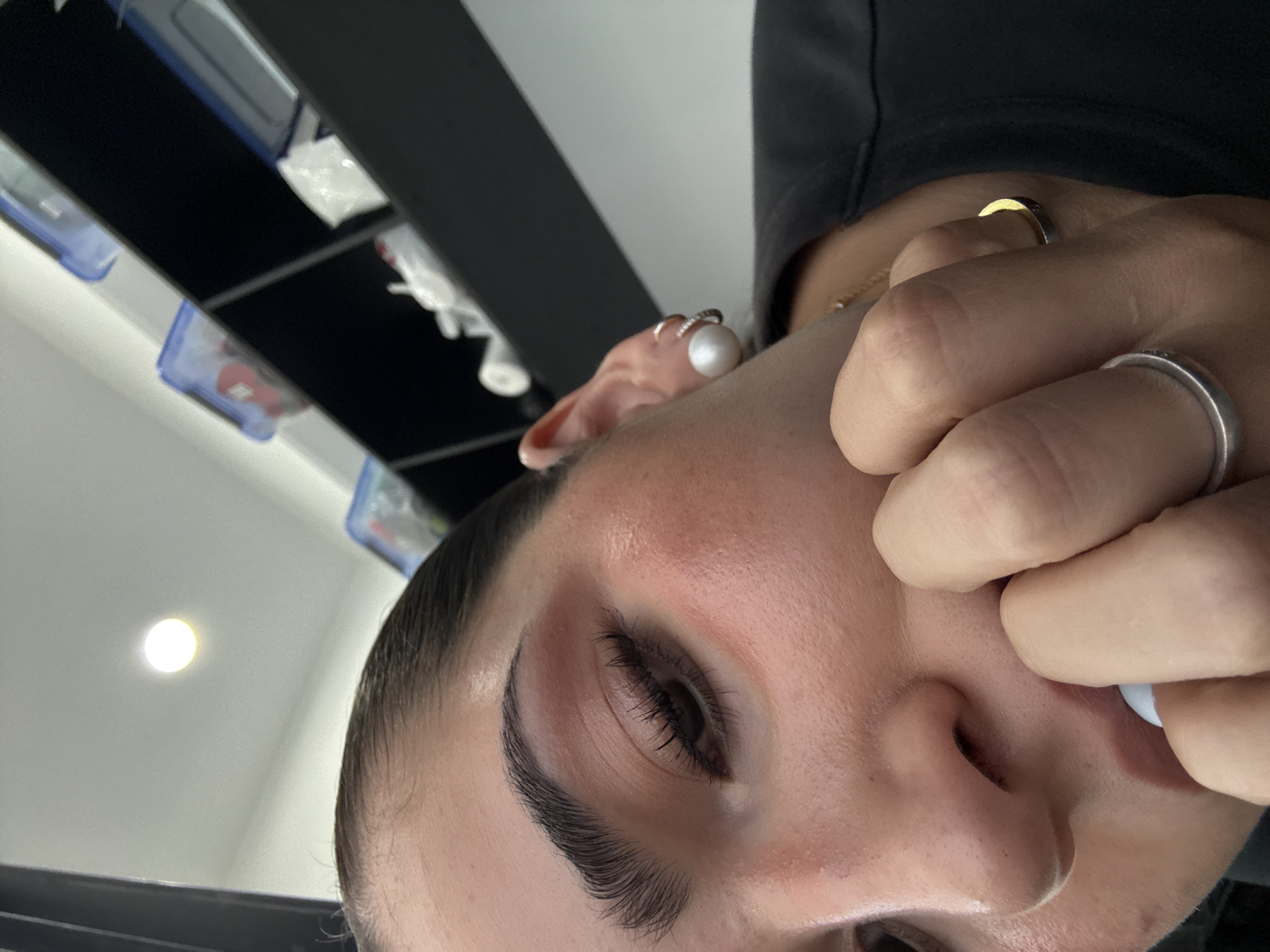 Close-up image of a person's face, likely a woman, with focus on the left eye and eyebrow. The person has dark eyebrows and eyelashes. The skin appears fair with some redness around the eye. The individual is wearing a pearl earring in their left ear and has a ring on their finger. Another hand is visible holding a white object near the person's mouth. The background shows a white ceiling with dark shelves containing various items. The location and city are not discernible from the image.