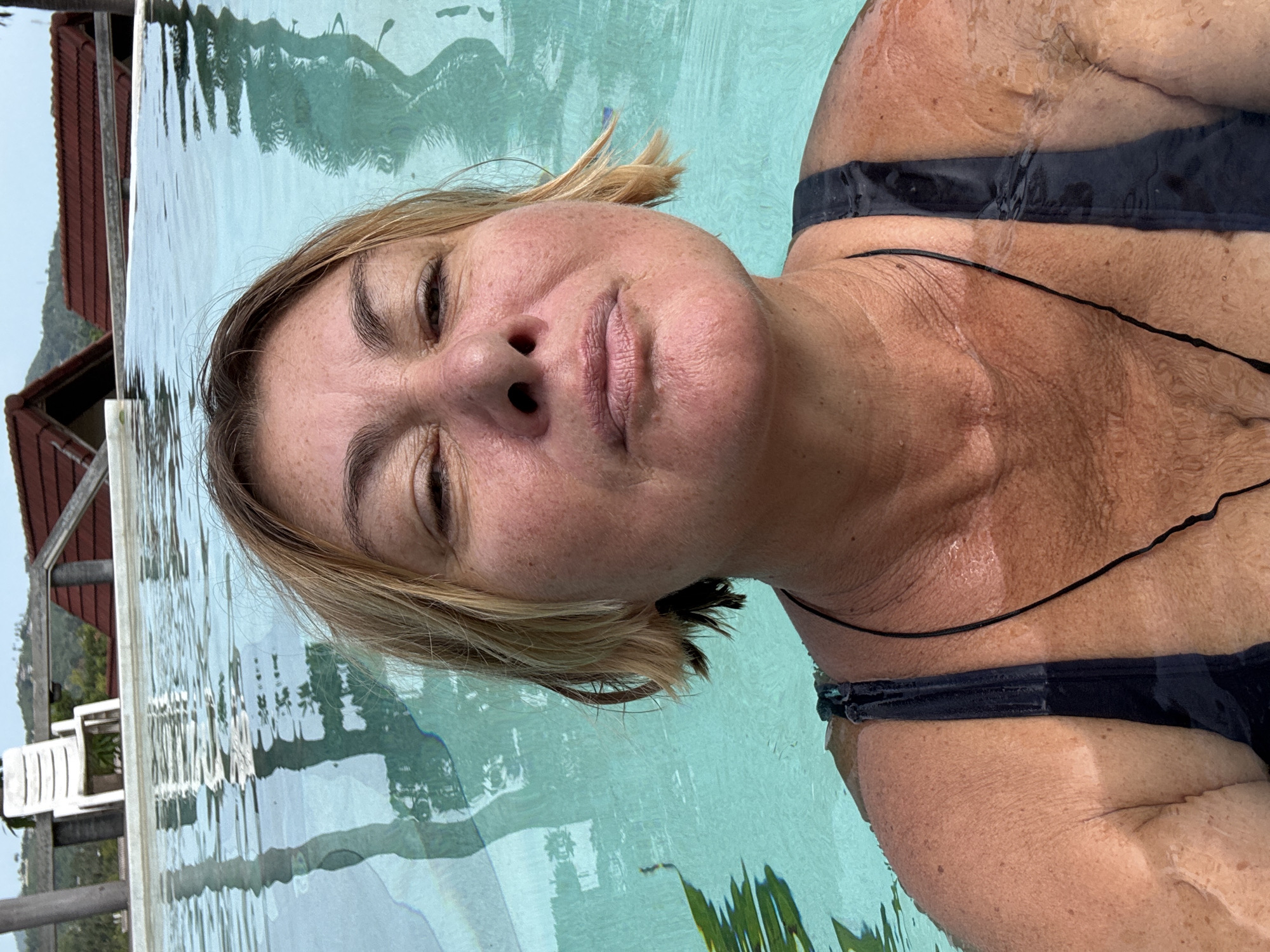 The image is a close-up shot of a woman immersed in a body of water, likely a swimming pool. She has fair skin and blonde hair, and is wearing a black swimsuit. The water is clear and turquoise in color. In the background, there is a glimpse of a building with a red roof, greenery, and some white patio furniture. Based on the location, it is likely that the image was taken in บ้านบางรักษ์, Thailand.