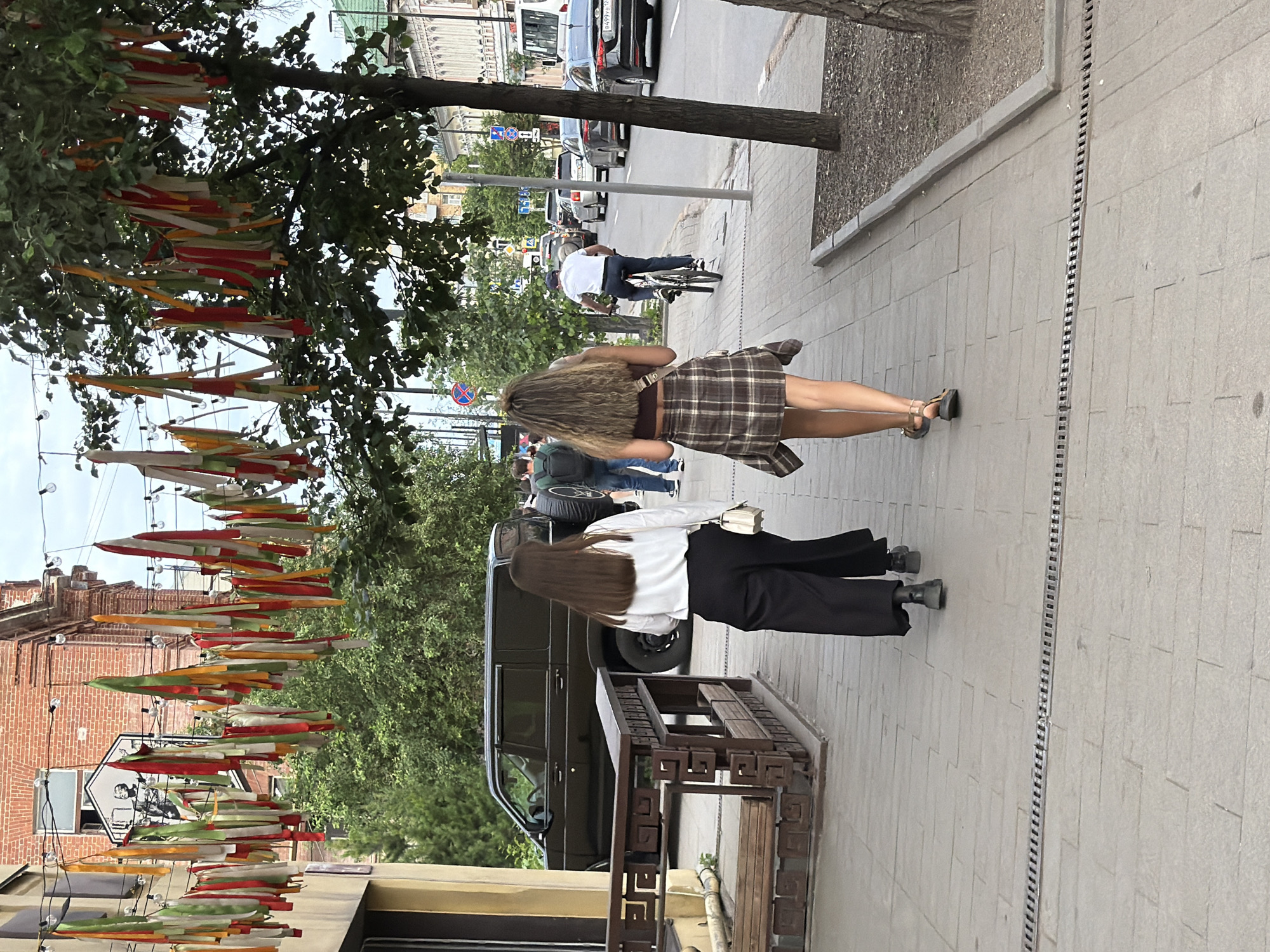 The image captures a street scene in Krasnoyarsk, Russia, likely during daytime. Two young women are walking away from the camera on a sidewalk. The woman on the left is wearing a white long-sleeved shirt and black wide-leg pants. The woman on the right has on a brown and white plaid shirt tied at the waist over a dark top and a plaid skirt, with sandals on her feet. A man on a bicycle is seen further down the sidewalk, cycling away from the camera. In the background, a dark-colored SUV is parked on the street, and several cars are driving on the road. The street is lined with trees and buildings. Decorative fabric streamers in red, white, green, and yellow are strung across the street overhead, adding a festive touch. The sidewalk is paved with light-colored rectangular tiles, and a drainage grate runs along its edge.