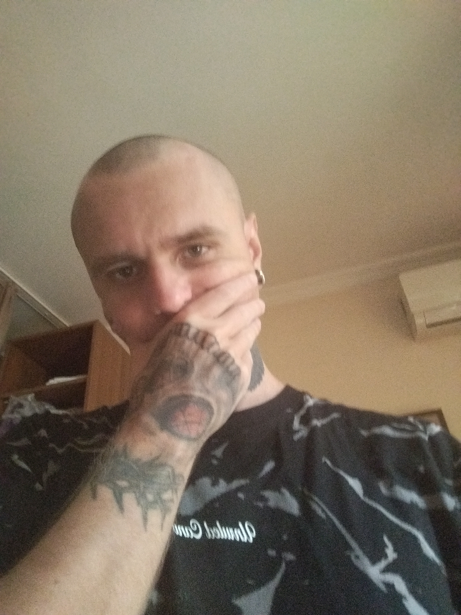The image is a selfie of a bald man with tattoos on his arm, covering his mouth with his hand. He is wearing a black t-shirt with a white, abstract pattern. The background appears to be an indoor environment, possibly a room in an apartment, with a light-colored ceiling and walls. There is a shelf unit with wooden shelves on the left side of the image, and an air conditioning unit mounted on the wall in the background on the right. The lighting suggests it may be daytime. The city of Batumi, Georgia, is not depicted in this image, nor is there any context provided that would place it there.