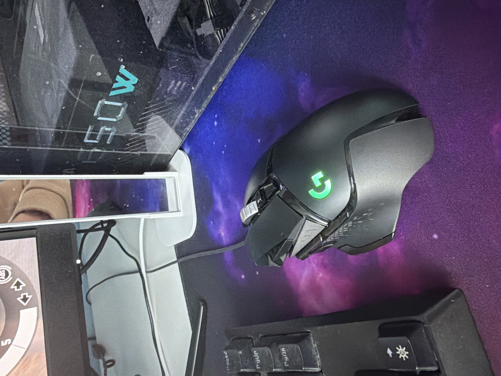 The image depicts a desk setup with a black Logitech G502 gaming mouse positioned on a purple galaxy-themed mousepad. Adjacent to the mouse is a black keyboard, partially visible in the lower right corner. A monitor displaying an interface with circular icons is located to the left. The monitor's stand is white, and there are visible cables and a white structure behind the monitor. The city and location are unknown.