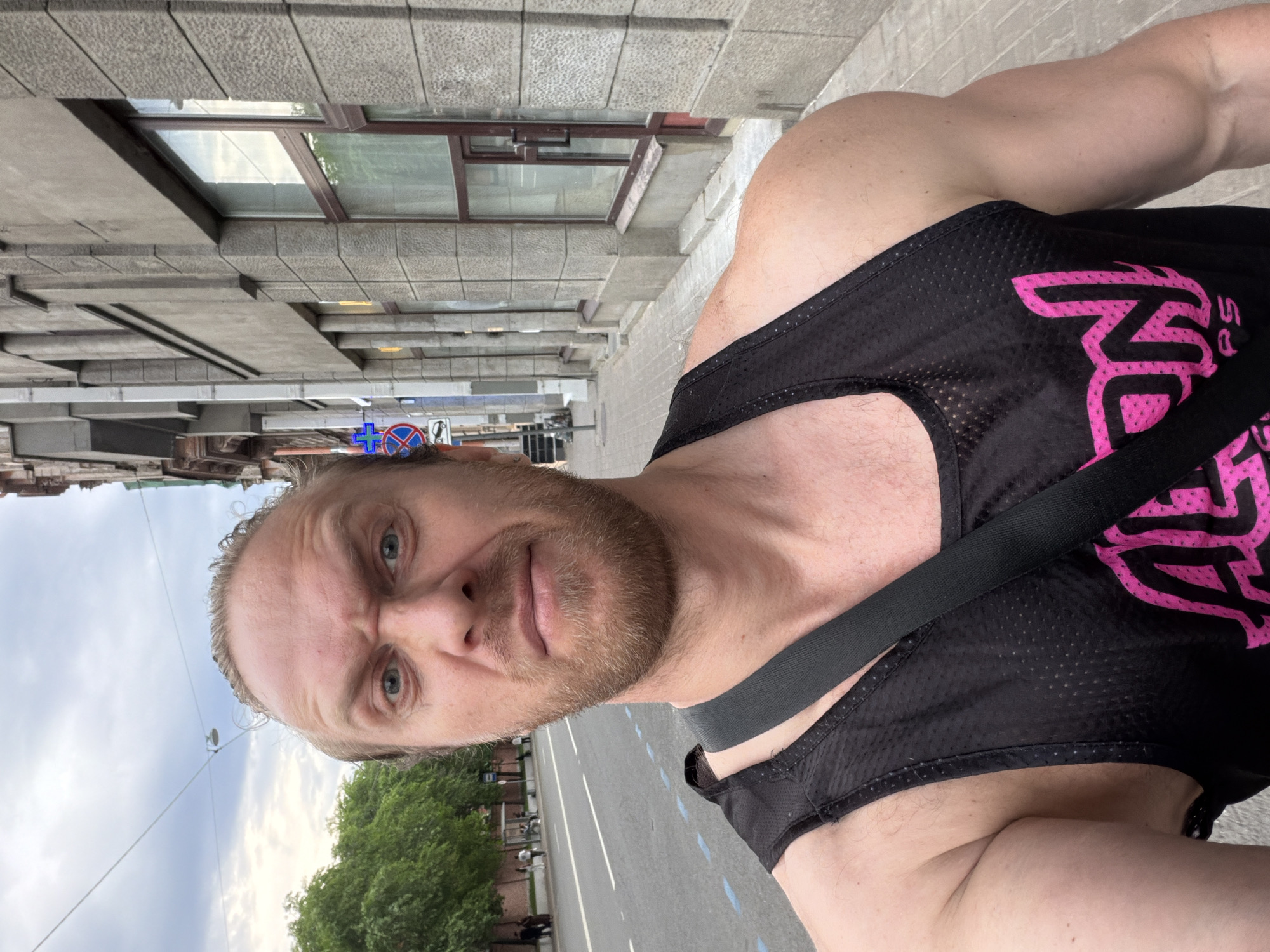 The image is a selfie featuring a muscular adult male subject outdoors on a street in Saint Petersburg, Russia. The subject occupies the lower half of the frame, looking directly into the camera with a strained or serious expression, indicated by furrowed brows and a slightly downturned mouth. He has light skin, blue eyes, a light-colored beard, and light-colored hair pulled back from his face. He wears a black mesh tank top, partially revealing a pink graphic logo on the right side of his chest, and a black strap crosses over his left shoulder. His prominent left arm and shoulder display visible muscle definition.
The background depicts an urban street scene. To the subject's left, a paved road with a solid double white line and dashed blue lines runs into the distance. A row of green-leafed trees lines the far side of the street, behind which several indistinct pedestrians are visible. The sky above is overcast with scattered clouds, and overhead wires are present. To the subject's right, a multi-story building with a grey, rough-textured stone or concrete facade dominates the background. The building features several rectangular windows with dark frames and is architecturally characterized by vertical pilasters, consistent with structures found in Saint Petersburg. A paved sidewalk runs alongside the building. Visible signs on a pole on the right include a blue circular sign with a white cross, a red circular 'no stopping' sign, and a white rectangular sign with a crossed-out car and 'P' indicating no parking.