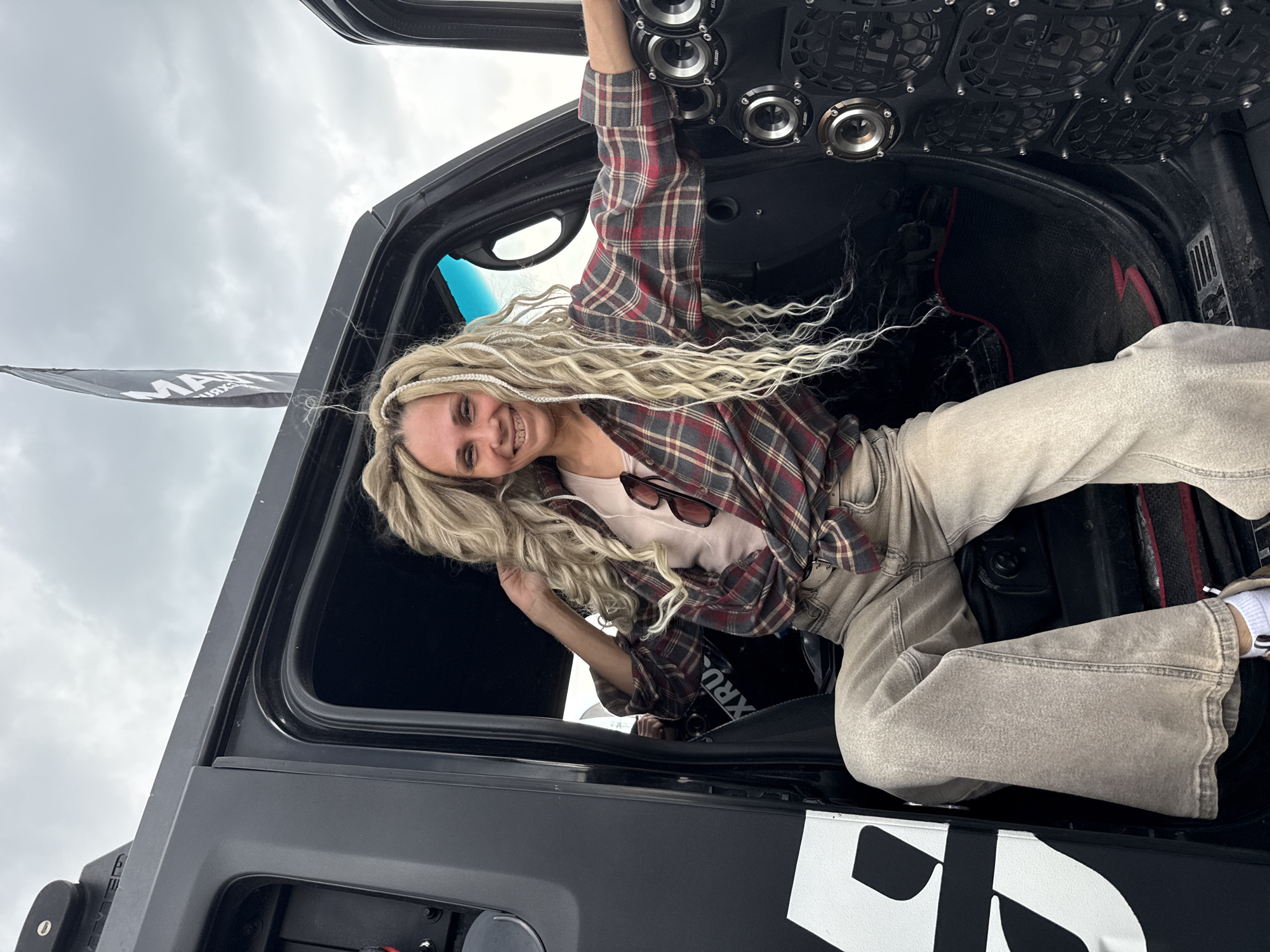 A woman is leaning out of a black vehicle. She has long blonde hair with braids and is wearing a plaid shirt, light-colored jeans, and white shoes. Sunglasses rest on her shirt. She is smiling. The interior of the vehicle has a speaker system overhead. Part of a flag is visible to the left. The image appears to be taken from below, looking up at the vehicle. The background is a cloudy sky. There is no visible evidence in the image that suggests it was taken in Rusanovo, Russia.