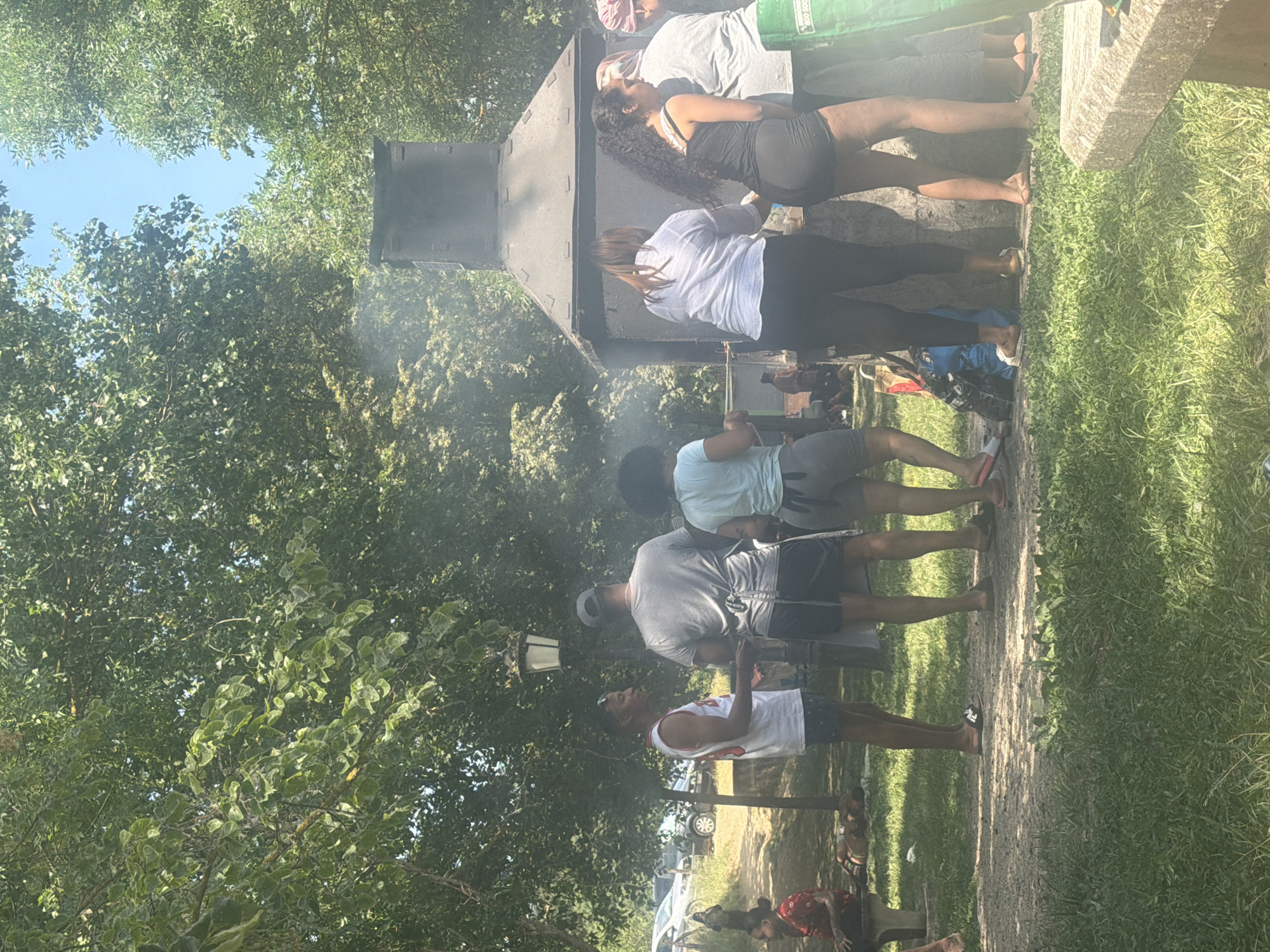 The image depicts an outdoor gathering, likely a barbecue or picnic, in a park-like setting with dense green trees overhead and a bright blue sky visible in patches. In the foreground, a group of people are gathered around a large, dark-colored barbecue structure that is emitting a significant amount of smoke. Several individuals are standing, some appearing to be tending to the grill, while others are observing or socializing. Clothing ranges from casual wear like t-shirts and shorts to more formal attire like a white blouse.
To the left of the barbecue area, a pathway or dirt track is visible, with several cars parked in the background. More trees and foliage line the pathway. In the immediate foreground and to the left, there are people sitting on a bench or the ground, some appearing to be resting.
The ground cover consists of both grass and a gravelly or dirt path. The overall atmosphere suggests a warm, sunny day, possibly in the summer. While the specific location of Campezo / Kanpezu, Spain is mentioned, the visual elements within the image do not provide any direct clues to confirm or deny this location. The scene captures a moment of communal activity and relaxation in an outdoor environment.