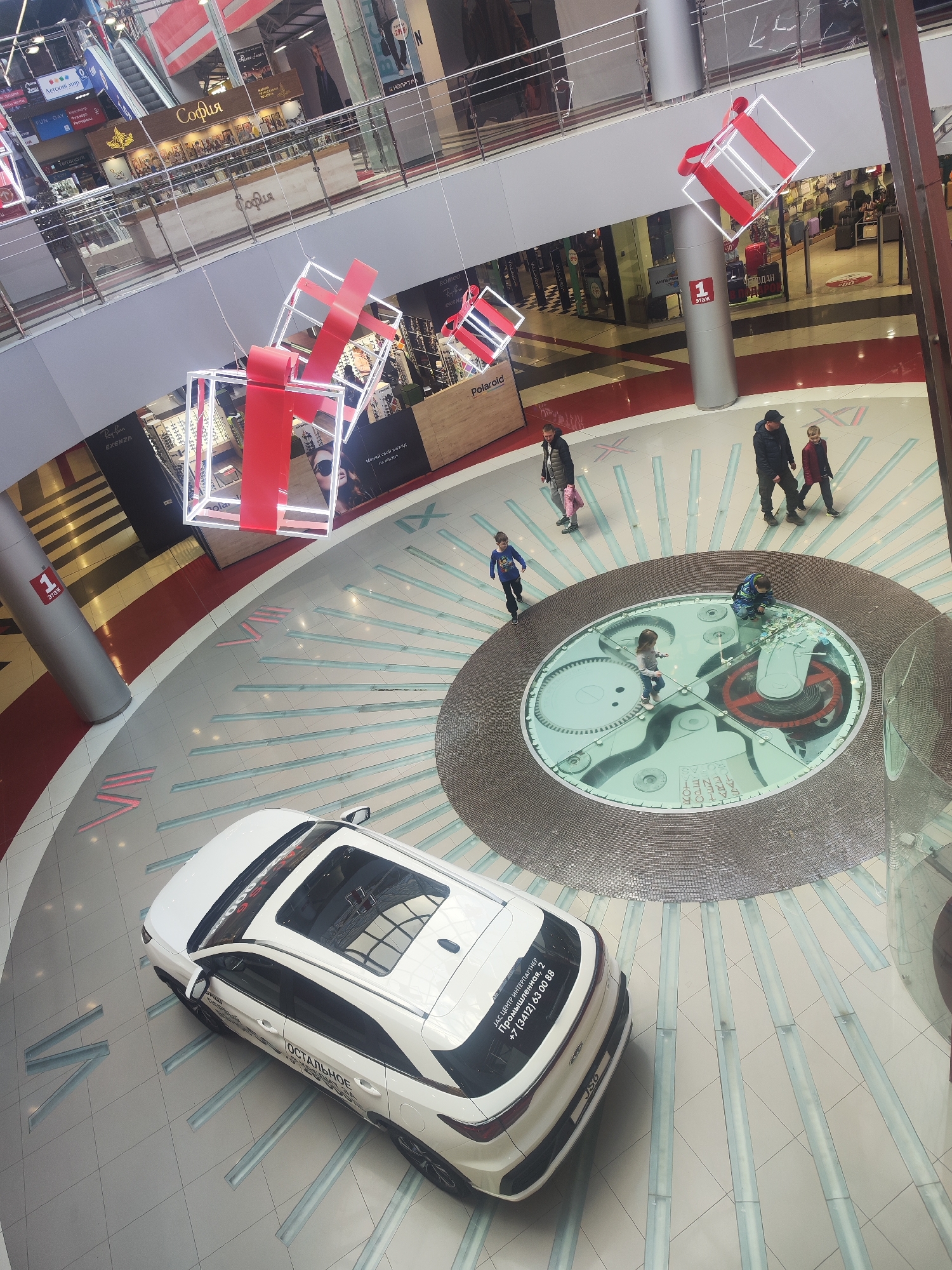 Here is a detailed description of the image:
**Overall Impression:** The image captures a high-angle view of an interior space, likely a shopping mall in Izhevsk, Russia. It prominently features a display car and a circular central area with a mechanical clock mechanism visible beneath a glass floor.
**Location:** Based on the sticker on the display car, it is likely taken in the "JAS CENTER INTERPARTNER" at "Промышленная, 2" in Izhevsk, Russia.
**People:** There are several people present in the image. Some are walking in groups, including an adult male and child. Several children are gathered around the glass floor with the clock mechanism, possibly observing or interacting with it.
**Subjects and Objects:**
* **White Car:** A white car with a sunroof is the most prominent object in the foreground. It has branding/stickers related to sales and service information.
* **Clock Mechanism Under Glass:** At the center of the image is a large, circular area featuring a transparent glass floor that reveals a complex mechanical clock mechanism underneath.
* **Decorative Elements:** Above the space, there are suspended decorative elements resembling gift boxes with red ribbon accents.
* **Storefronts:** The image captures glimpses of storefronts from brands such as Ray-Ban, Exenza, Rime Jeans, Polaroid, and София.
* **Columns:** There are supporting columns wrapped with branding and signage, including a floor number.
**Actions and Interactions:**
* People are walking around and exploring the area, including interacting with the car.
* Children are observing the clock mechanism under the glass floor.
**Environment:**
* The floor is tiled and marked with radial lines emanating from the center, possibly mimicking the appearance of a clock face.
* The space is well-lit, and the surrounding environment appears to be a modern and upscale shopping mall.
* The decorations and architecture suggest the space is a commercial environment designed to attract shoppers.
**Lighting:**
The scene is well-lit with overhead lighting, highlighting the various features and providing good visibility.