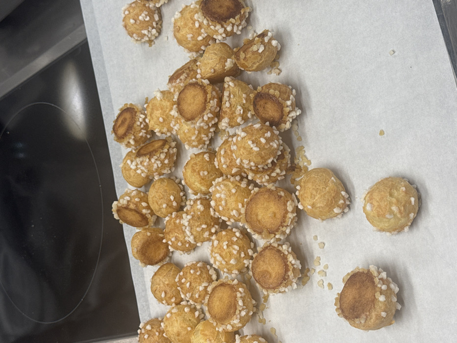 The image displays approximately 60 small, spherical golden-brown pastry items, consistent with *choux* puffs or *profiteroles*, arranged on a white sheet of parchment paper. Most of these pastries are adorned with a darker, flatter disc-shaped topping, suggestive of *craquelin*, and heavily sprinkled with irregular white pearl sugar, which is also scattered loosely across the parchment surface. The parchment paper rests on a dark, reflective surface, identifying as a stovetop or similar kitchen counter, with two faint circular hob outlines visible in the blurred background. No individuals are present in the scene. The specific location within Paris, France, cannot be determined from the visual information provided, as the environment is a generic indoor kitchen setting.