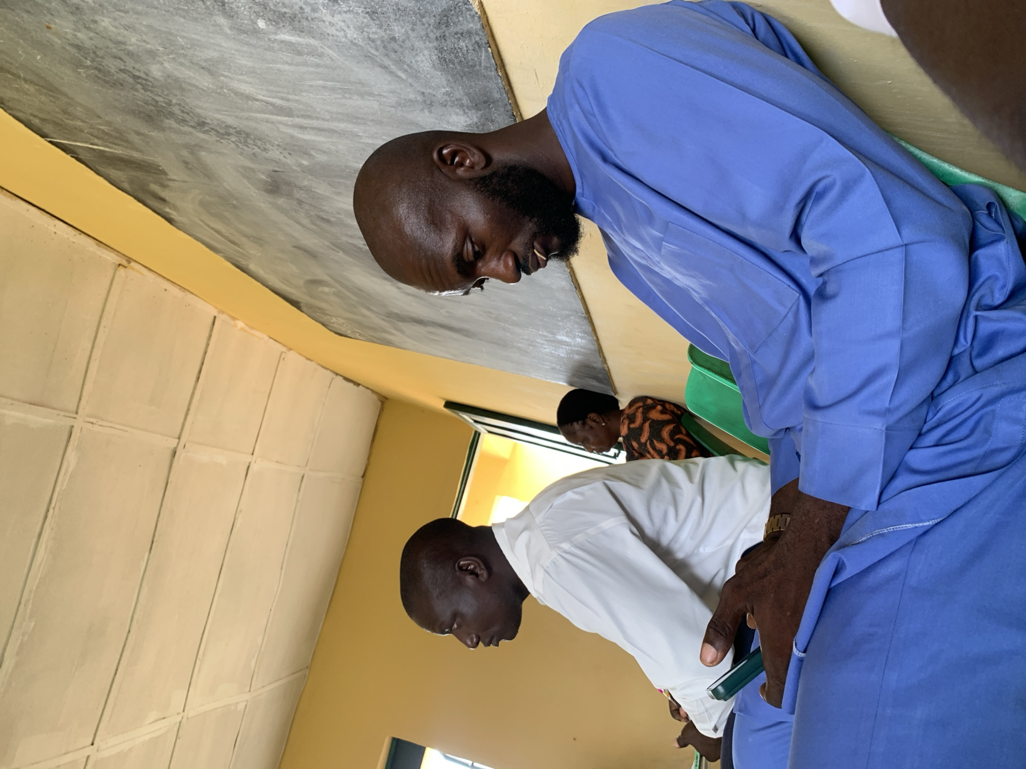 This rotated image, taken in Akwanga, Nigeria, depicts an indoor scene featuring three individuals, two men in the foreground and one woman in the background, all seated.
The prominent figure in the right foreground is a man with a bald head, a dark beard, and glasses perched on his nose. He is wearing a bright blue long-sleeved tunic (likely a kaftan or similar traditional attire) and appears to be looking down with a focused expression. His right hand, which has a gold-toned watch on the wrist, holds a dark green object, possibly a smartphone or a small book. He is seated on a green chair, a portion of which is visible under his arm.
To his left, slightly further back, is another man with a bald head, dressed in a white long-sleeved collared shirt. He is also looking downwards, mirroring the focused posture of the first man.
In the distant background, a woman is partially visible. She is wearing a patterned top, possibly in shades of brown and orange, and is also seated, facing in the same general direction as the men.
The setting appears to be a modest room, possibly a classroom or meeting area. The walls are painted a light yellow or cream color. The ceiling is visible across the top of the frame; it shows a mixture of greyish, mottled textures and areas with white rectangular or square panels arranged in a grid pattern. A doorway or window opening is discernible in the background, allowing some light into the room. The overall impression is one of quiet contemplation or attention to an activity not fully visible within the frame. There is no clearly legible text in the image. The time of day is not explicitly indicated but seems to be daylight hours.
