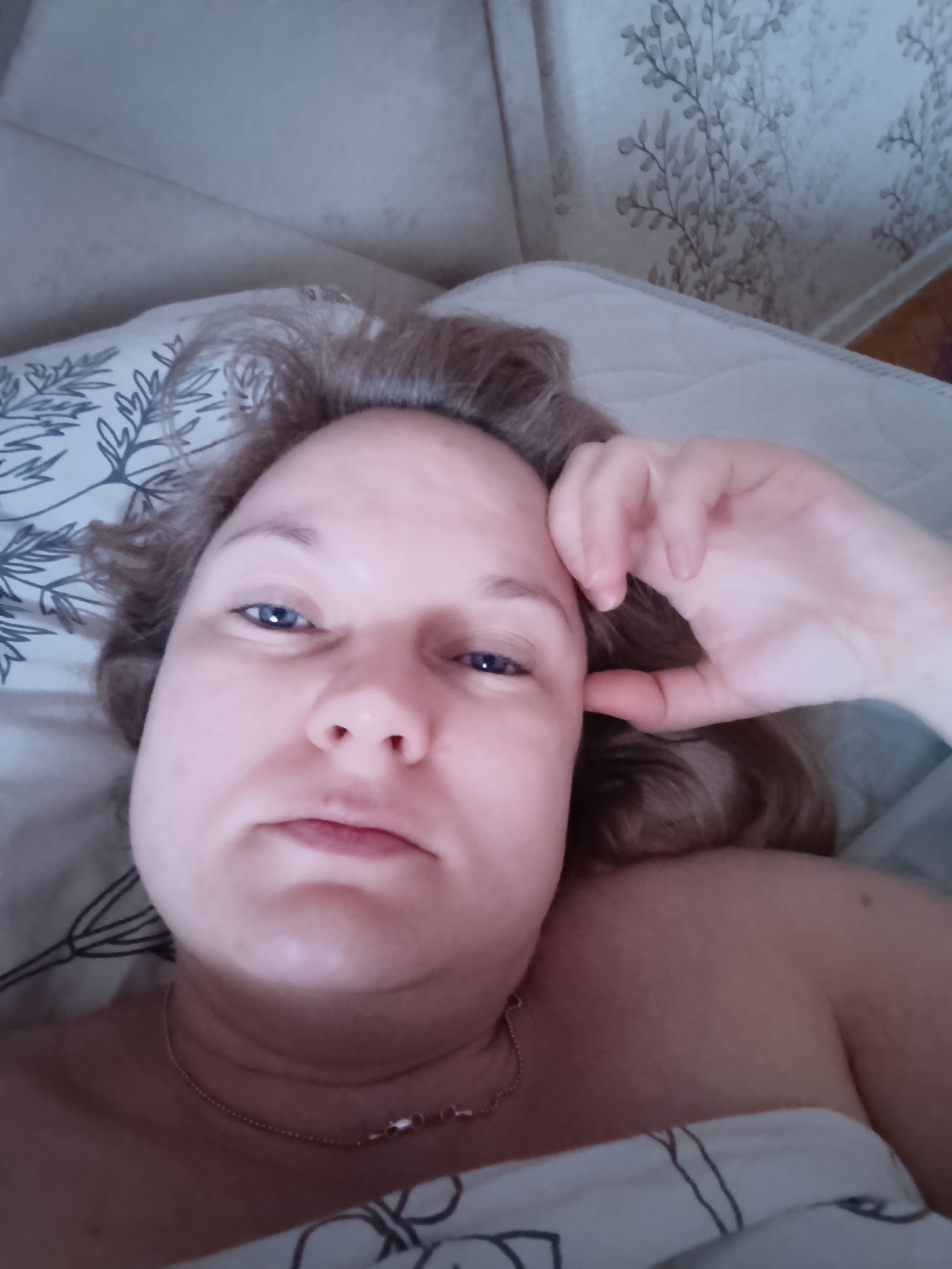 A woman with fair skin and blue eyes is lying in bed. She is wearing a gold necklace. Her hair is light brown. She is holding her hand up to her face. The background is a patterned wall and bedding. The location is unknown.