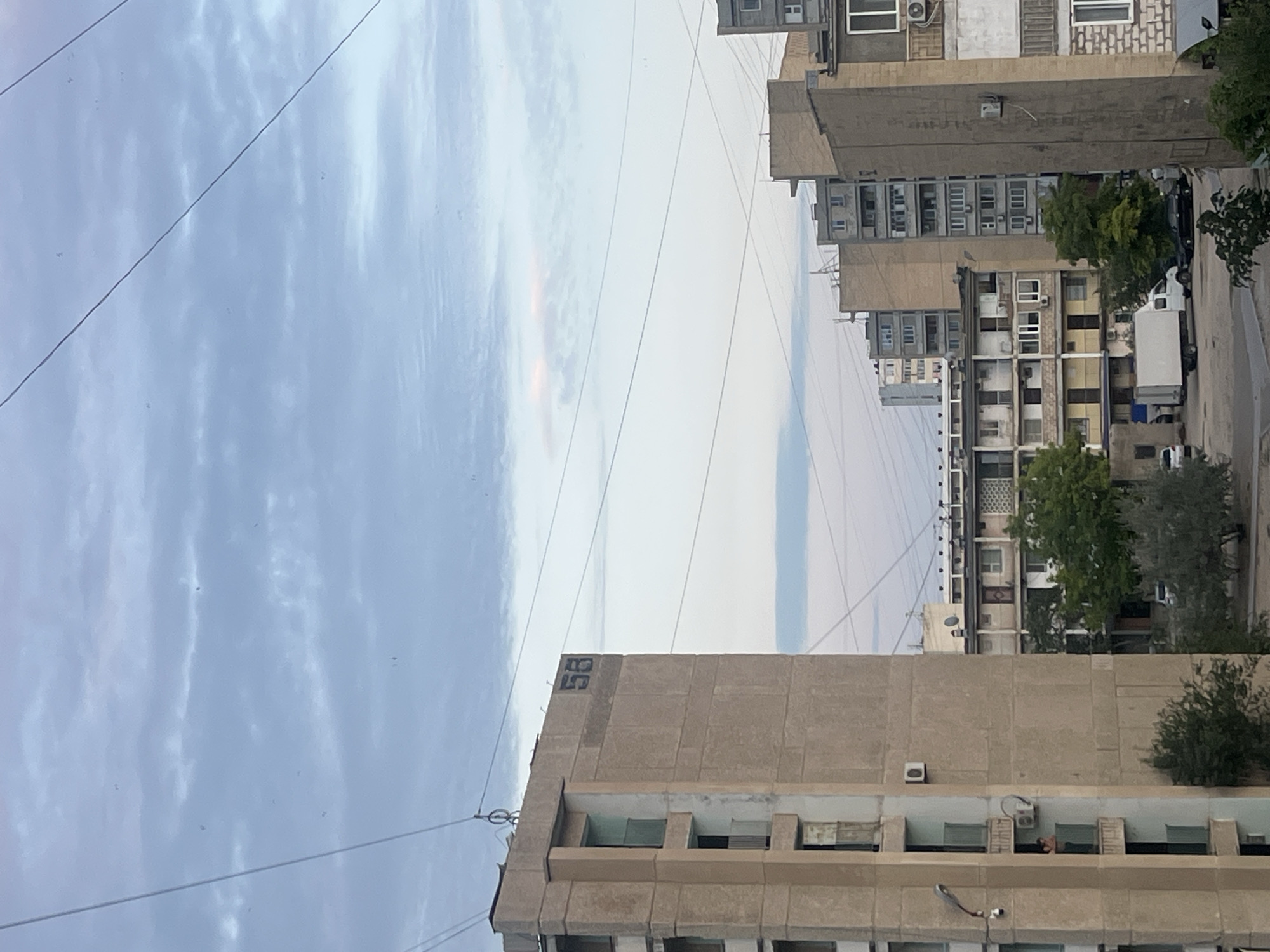 This image depicts a view of apartment buildings in Aktau, Kazakhstan, under a cloudy sky. The architecture suggests a Soviet-era housing block style, characterized by concrete facades and balconies. In the foreground, a tall, beige apartment building with visible balconies and air conditioning units is prominent. The number "58" is spray-painted in black on the upper facade of this building. Further into the scene, other apartment buildings of varying heights and architectural styles are visible, including one with a patterned facade. In the lower right, a partial view of another building shows a brick facade. The street level includes parked vehicles, a white truck, and some trees. Power lines crisscross the sky, and a few small, dark specks, possibly birds, are present in the sky. One person is visible on a balcony of the foreground building, leaning over the railing. The lighting suggests it is either early morning or late afternoon.