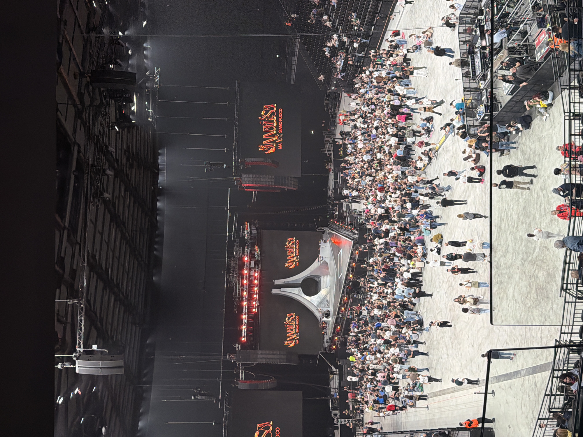 The image shows a concert audience gathered in a large, indoor venue, likely the Forum di Assago in Milan, Italy, based on the presence of Italian text on the stage screens. The crowd is dense in front of the stage, with seating visible in the upper tiers. A large stage setup includes screens displaying "Siamofuoco" in red, illuminated architectural structures, and lighting rigs. People are standing and milling about on the floor area in front of the stage. Construction and support structure for the roof of the venue are clearly visible.