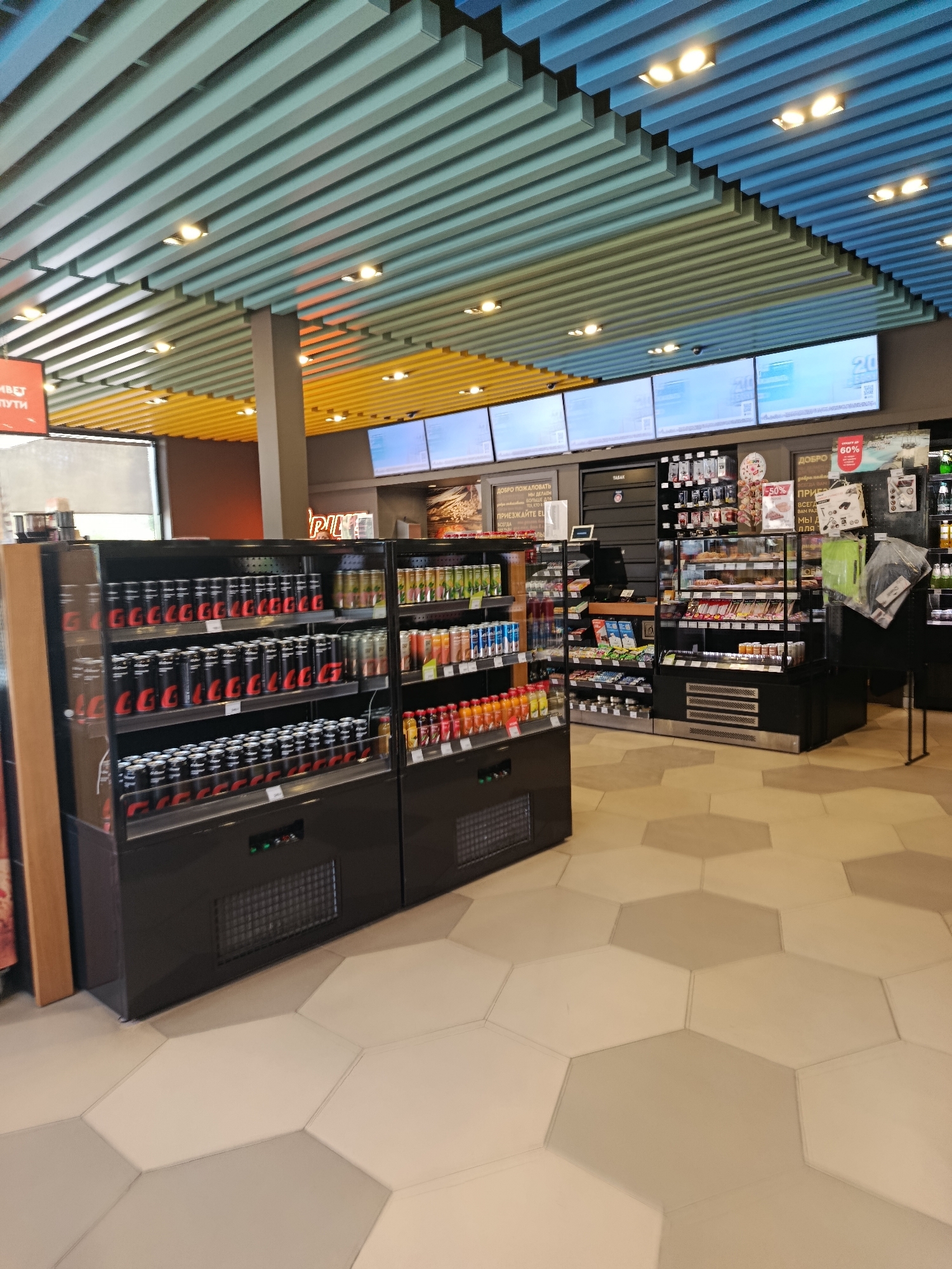 The image depicts the interior of a convenience store, possibly a gas station convenience store, given the branding and product selection visible. The ceiling features a modern design with slatted panels in blue and yellow, interspersed with track lighting. Large digital screens are mounted on the wall above a counter area.
In the foreground, to the left, is a refrigerated display case filled with rows of black cans, many with red branding. To the right of this is another display case containing various bottled and canned beverages. Further to the right, a glass-doored display case shows an assortment of food items, including what appear to be pastries and pre-packaged sandwiches. Behind these, shelves are stocked with more packaged goods and a display of tobacco products.
The floor is tiled in a hexagonal pattern, alternating between light beige and grey. The overall environment suggests a retail space designed for quick purchases, with a variety of refreshments and convenience items. There are no people visible in the image. The location is identified as Orekhovo-Zuyevo, Russia.