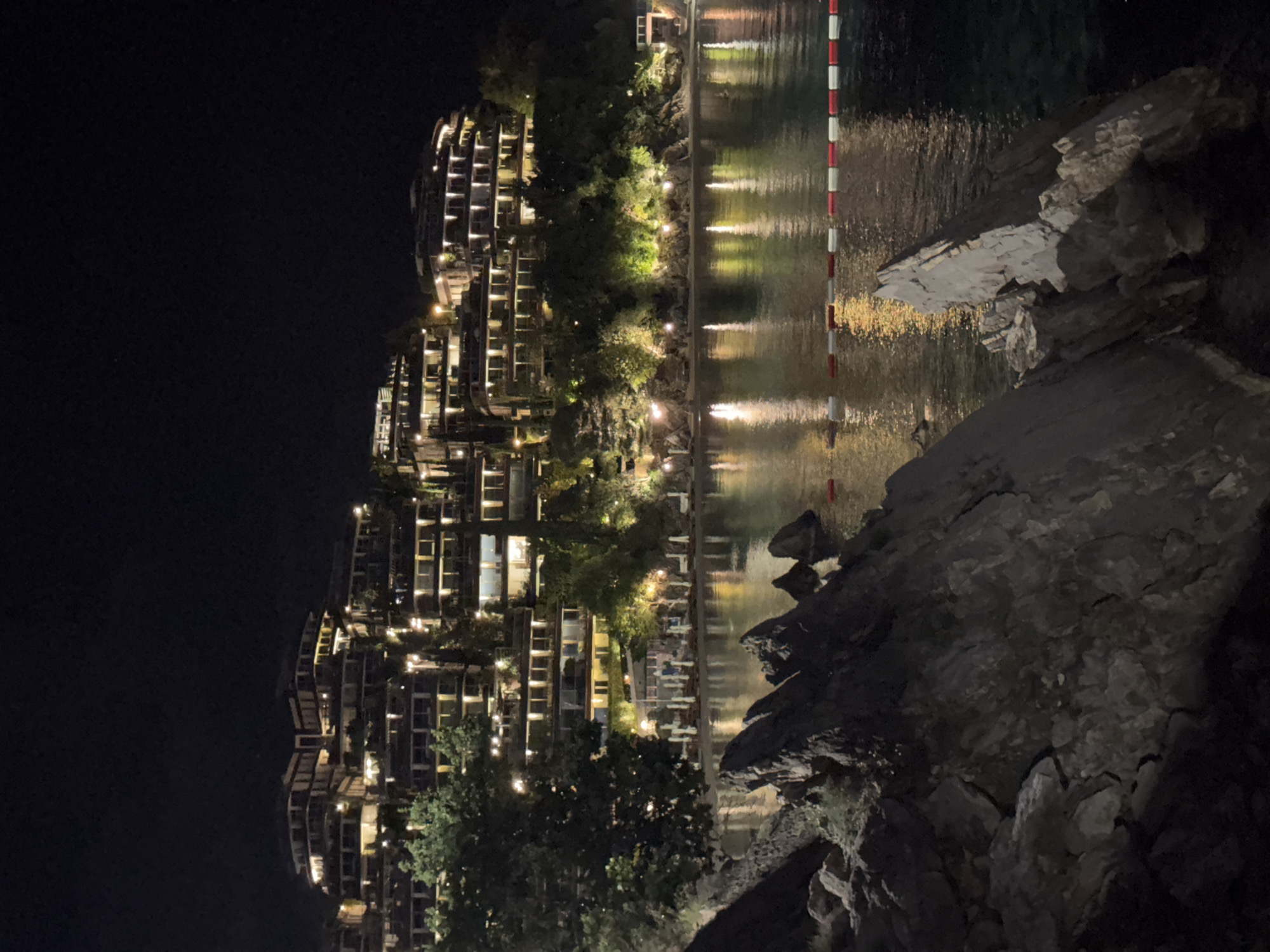 The image depicts a nighttime view of a waterfront scene. In the background, there is a multi-story building complex with balconies lit up with lights, suggesting residential or resort use. The building is positioned on a slope. The building is located in Budva, Montenegro. In the mid-ground, the water reflects the lights from the building and nearby areas. A vertical striped pole is visible near the water's edge. In the foreground, there is a rocky area, possibly a shoreline or cliff, with dark-colored rocks and some vegetation.