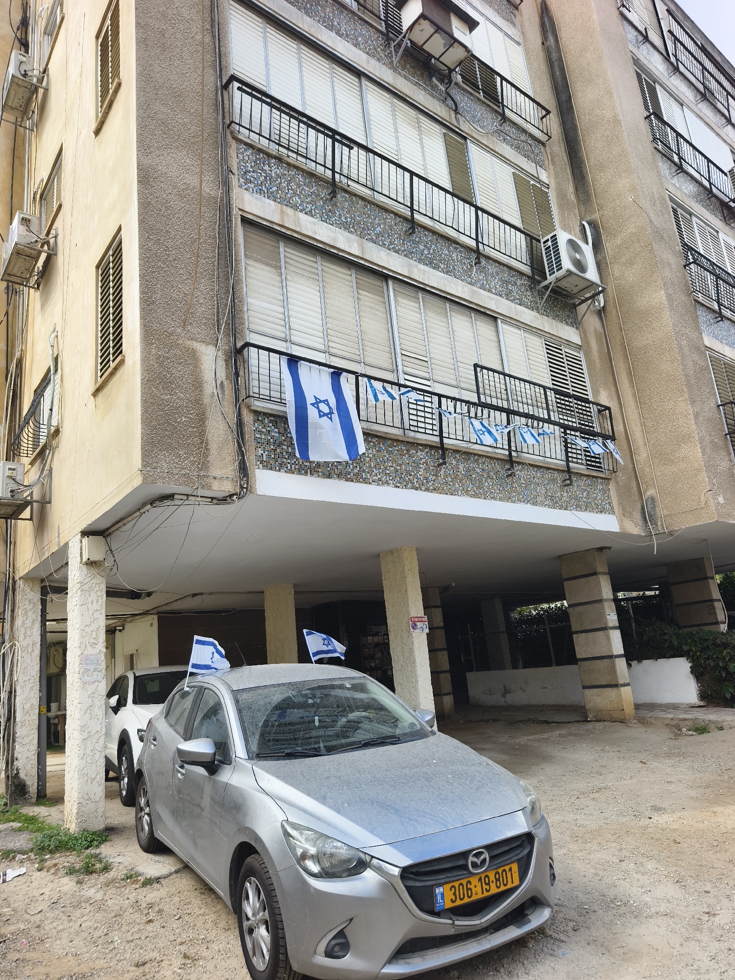 The image shows a multi-story building in Petah Tikva, Israel, decorated with Israeli flags hanging from a balcony. A silver Mazda car with license plate "306-19-801" is parked in front of the building, also adorned with an Israeli flag. A white car is parked behind the Mazda. The building appears to be an apartment complex with balconies and external air conditioning units. The area in front of the building is unpaved.