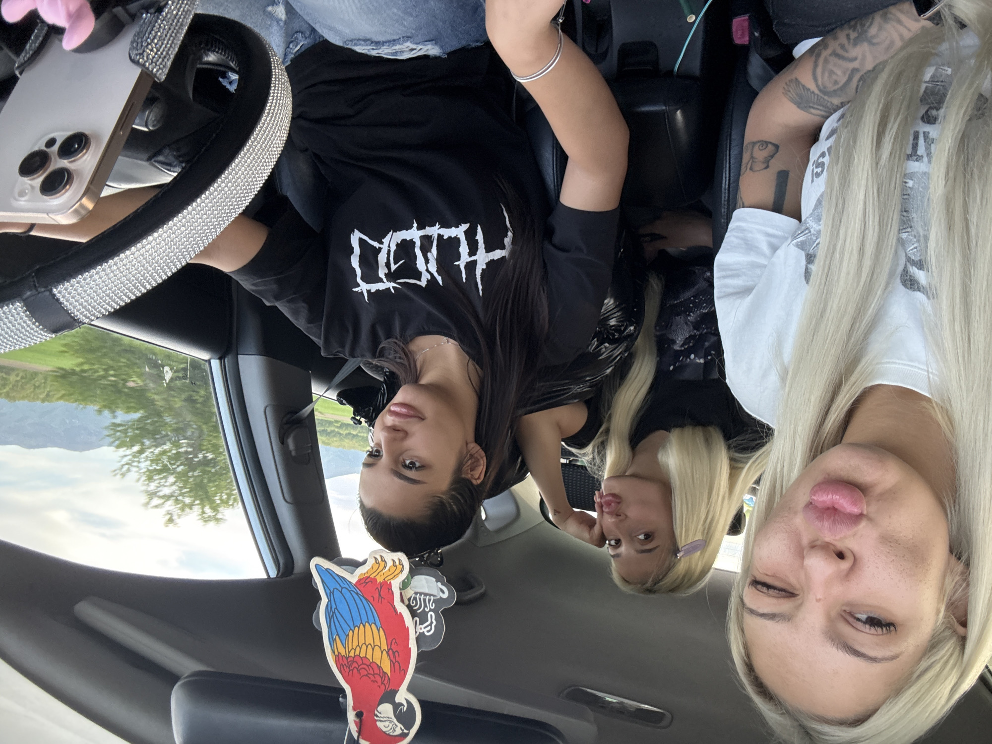 Three women are in a car, presumably parked, based on the scenery visible through the car windows. The woman on the left has dark hair and is wearing a black t-shirt with white lettering. The two women on the right have blonde hair. The woman in the middle is partially visible, and the woman on the far right is winking at the camera. The steering wheel is covered in rhinestones, and a phone is mounted next to it. A parrot air freshener hangs from the rearview mirror. The location appears to be Tulovo, Bulgaria, suggested by the surrounding landscape of rolling hills and sparse trees.