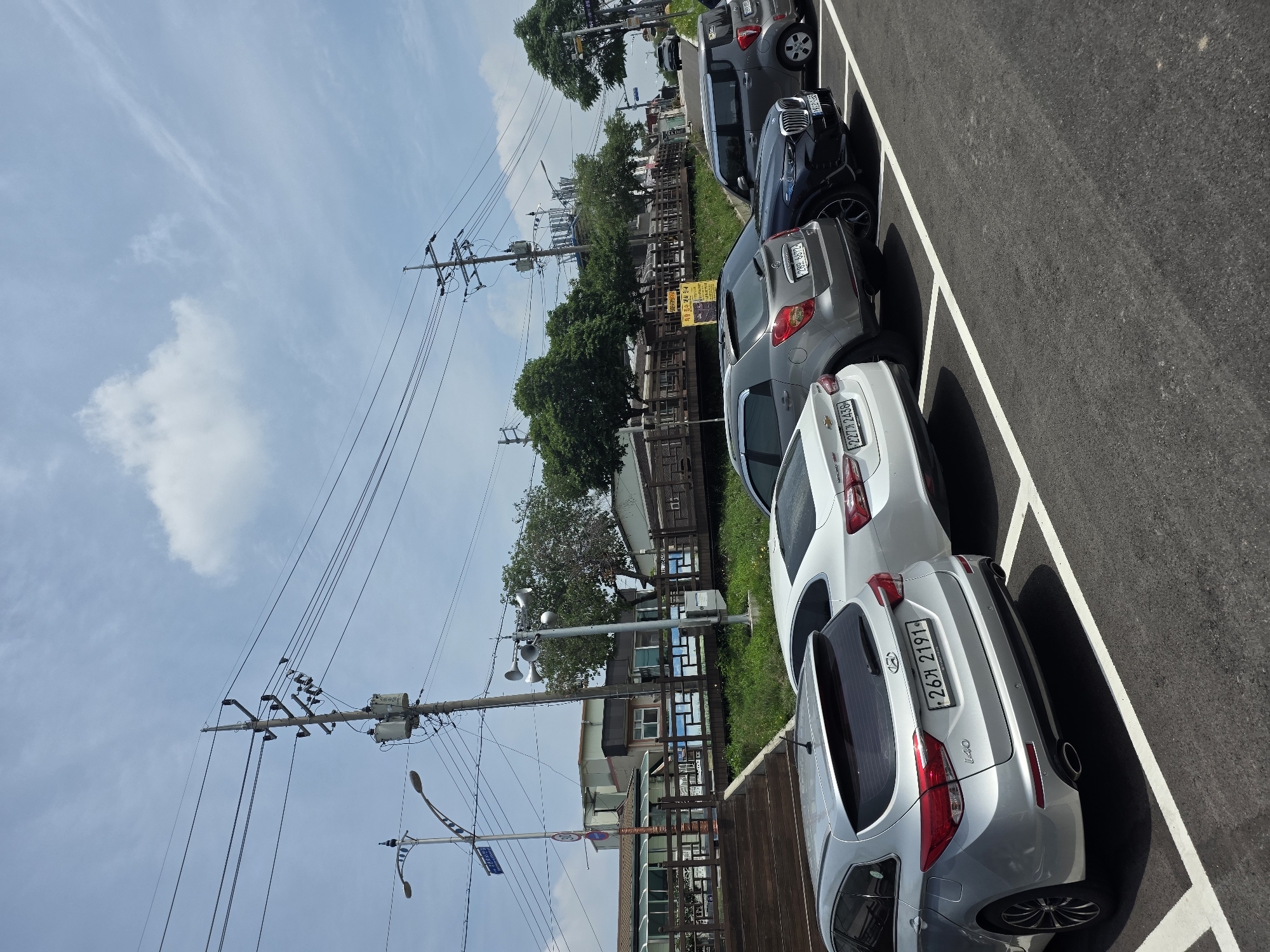 This is a daylight, eye-level, outdoor, medium shot of a parking lot in Anseong-si, South Korea. Six cars are parked in a row on an asphalt surface marked with white parking lines. The cars are, from left to right: a silver Hyundai i40 wagon, a white Chevrolet Malibu, a gray SsangYong Tivoli, a black BMW X6, a gray Kia Morning, and a white Hyundai Avante. In the background, there is a wooden deck with stairs leading up to a building with a green roof and glass windows. Beyond the building, trees and utility poles with power lines are visible against a bright blue sky with scattered clouds. A street sign is visible on a pole to the left of the wooden deck. The overall scene depicts a typical urban parking area with surrounding infrastructure under a daytime sky.