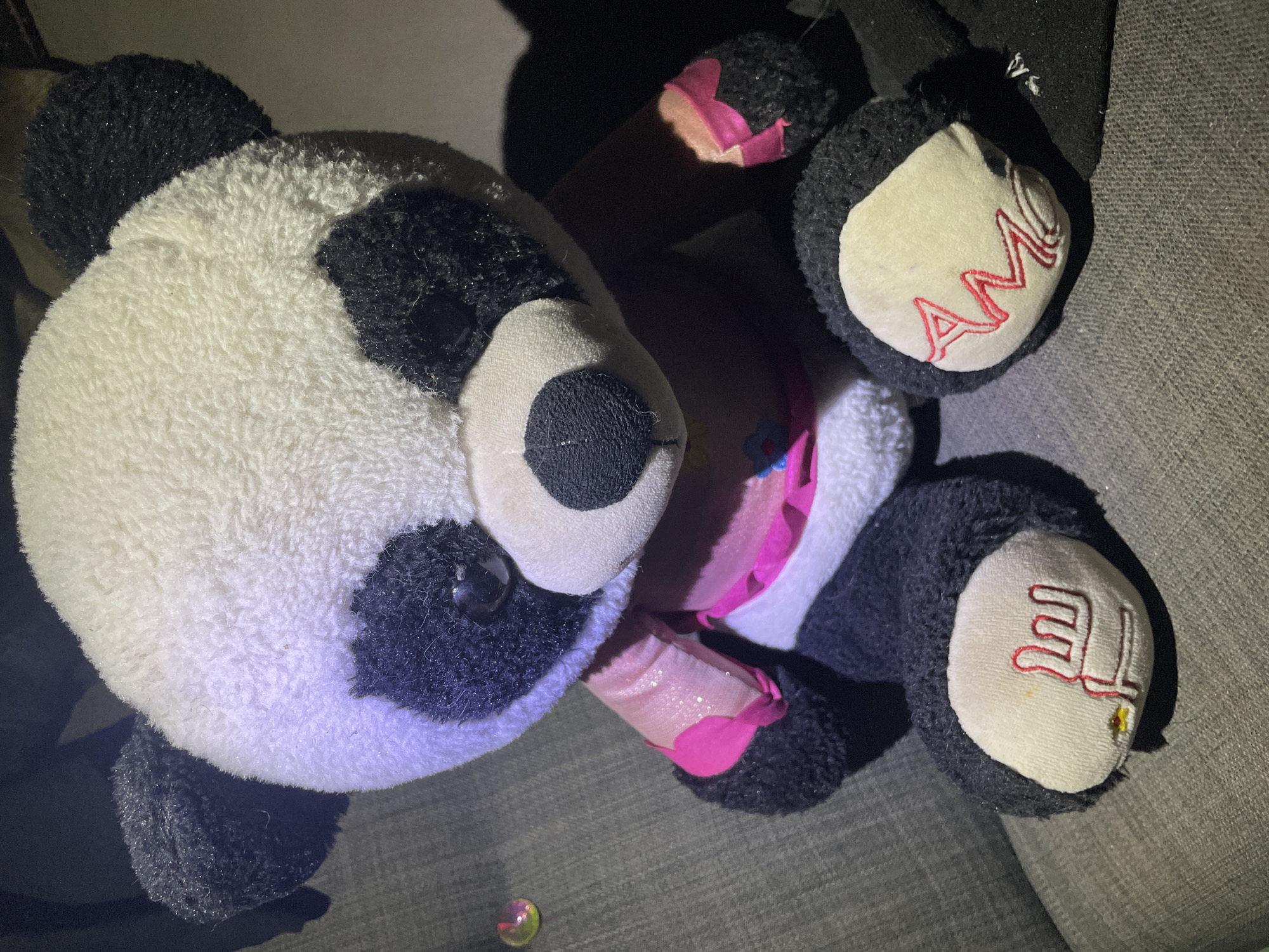A stuffed panda bear sits on a gray couch. The panda is white with black ears, eye patches, and limbs. It wears a pink and sparkly outfit. There are red embroidered designs on its feet. A small, colorful bead is visible to the left of the bear. The lighting is dim, with a spotlight creating a purple hue on the bear's head and casting shadows. There is no indication of any specific location within Iyafin, Nigeria.