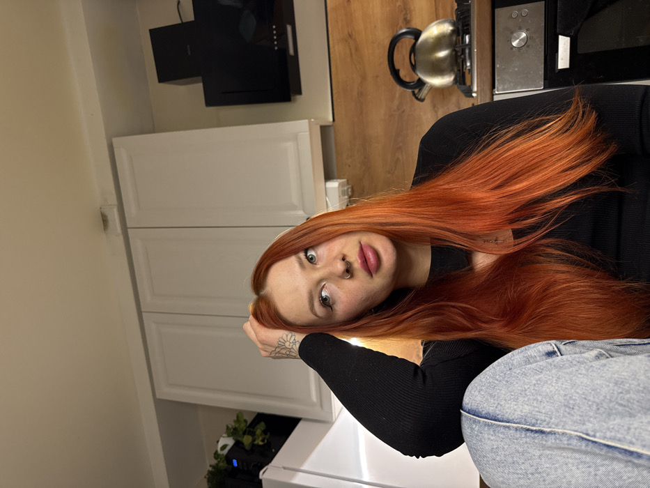 A young woman with long, vibrant, reddish-orange hair is captured in a close-up, rotated portrait. Her hair, which appears to be straight and shiny, cascades over her shoulders and chest, partially obscuring her right arm. She has a fair complexion, wide, light-colored eyes that gaze directly at the viewer, and dark eyebrows. Her lips are full with a soft pink hue, and she has a septum piercing and another small piercing on her right nostril. A tattoo, possibly a mandala or floral design, is visible on the back of her left hand or wrist.
She is wearing a black, ribbed, long-sleeved top. Part of her lower body is visible on the right, showing light blue denim jeans with a slightly distressed or faded appearance around the knee area. Her left arm is bent, with her hand positioned near her head.
The background suggests an indoor domestic setting, likely a kitchen or utility area. Behind her, to the left, are white kitchen cabinets with panel doors. Above the cabinets and further back, a modern black range hood is mounted on the wall. A wooden-look countertop extends across the middle ground. On the countertop to the woman's right, a shiny, metallic kettle is visible, along with part of a silver-colored appliance, possibly a microwave or oven, featuring control knobs and a digital display. A white electrical outlet or switch is also present on the wall above the countertop. On a lower surface to the left, below the white cabinets, some green potted plants are visible.
The overall image is rotated 90 degrees clockwise. The woman's expression is direct and engaging. There is no visible text in the image.