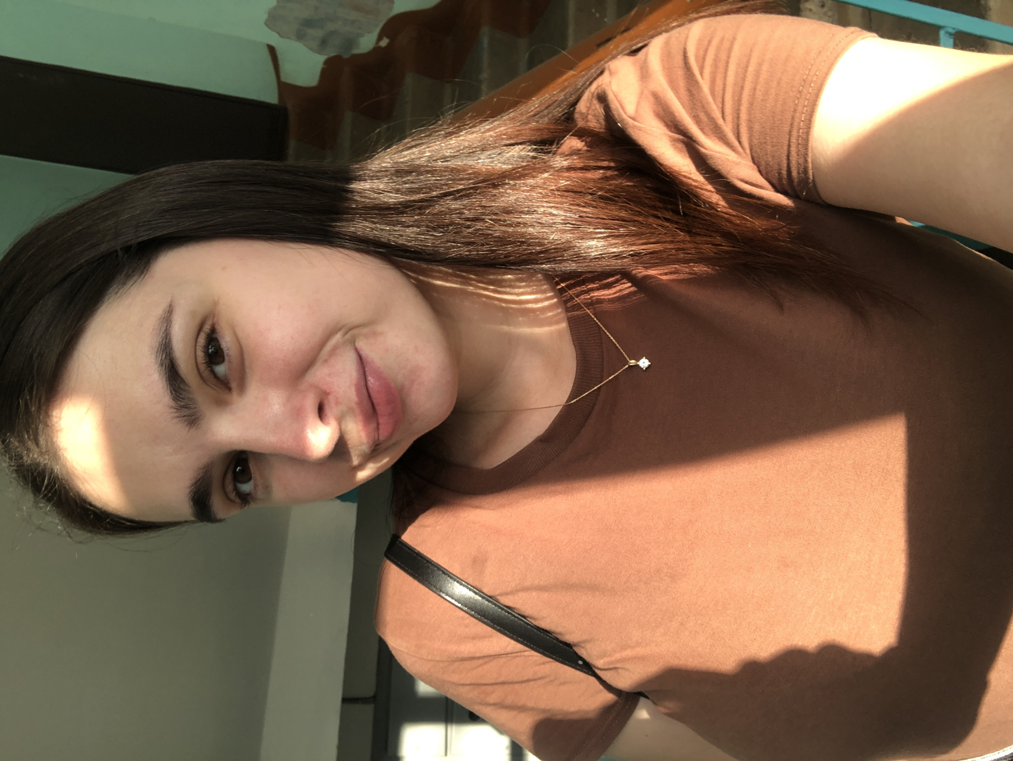 A young woman with long, dark brown hair, wearing a brown t-shirt and a thin gold chain with a diamond pendant, looks into the camera. She has a light complexion and brown eyes. A strap from a black bag is visible over her left shoulder. The lighting suggests she is indoors, near a window, with sunlight casting shadows on her face and clothing. The background is blurred, showing a wall with some textured patterns and possibly a staircase. The environment appears to be a residential building or an indoor public space. The image was taken in Chaikovsky, Russia.