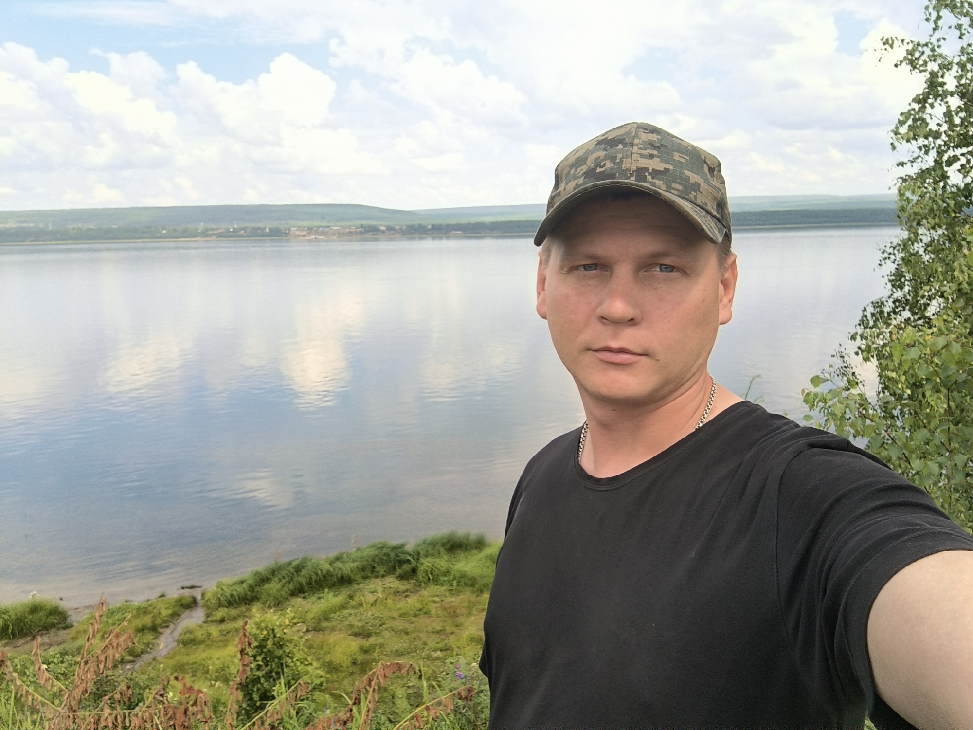 A man in a camouflage baseball cap and a black t-shirt is taking a selfie outdoors. He is positioned on the right side of the frame, looking directly at the camera. Behind him, a large body of water, likely a river or lake, reflects the sky and clouds. The water stretches towards a distant, tree-covered shoreline under a cloudy sky. The foreground features green grass and some dry, brown plants. The man appears to be standing on a slight incline overlooking the water. The setting suggests a natural environment, possibly a park or scenic overlook within or near the city of Boguchany, Russia.