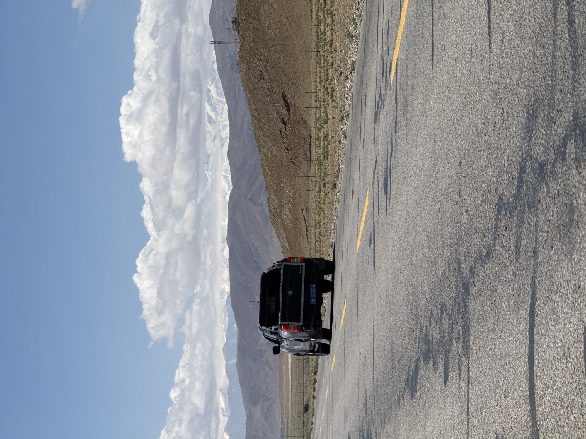 The image captures a black pickup truck, identified as a Dongfeng Rich, parked on the shoulder of a paved road in a vast mountainous landscape. The road features clear yellow markings. Bright daylight is evident from the long, distinct shadows cast across the asphalt.
In the background, rugged mountains dominate the horizon, with several peaks exhibiting patches of snow, suggesting a high altitude. The hillsides are largely barren and brown, interspersed with sparse green vegetation and a fence running parallel to the road. Distant transmission towers are visible on one of the mountain slopes. The sky above is a clear, vibrant blue, dotted with large, fluffy white clouds.
The truck's rear is visible, displaying the license plate "甘B 35566". No people are present in the scene. The setting is Mainpu township, China, and the overall atmosphere suggests a serene and expansive natural environment, typical of a remote road.