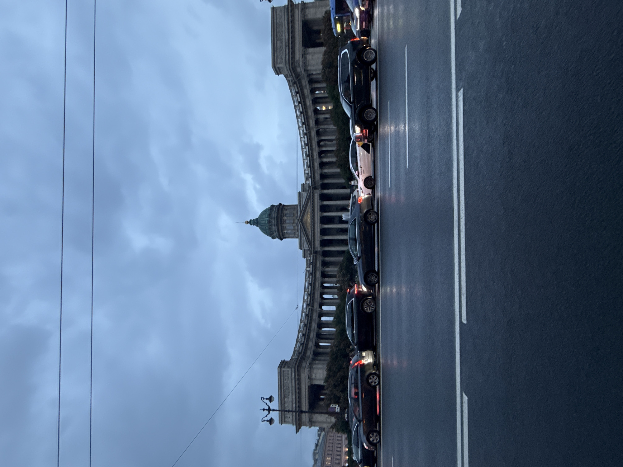 The image shows a view of Kazan Cathedral on Nevsky Prospekt in Saint Petersburg, Russia. The sky is overcast and a deep blue, with thin, dark clouds. Several cars are stopped in traffic on the wet street in front of the cathedral, which is a large, neoclassical building with a prominent dome and colonnade. The street has white lane markings and reflections of lights from the cars can be seen on the wet surface. The overall mood of the image is somber and muted, suggesting it was taken during twilight or on a heavily clouded day. No people are clearly visible or identifiable.