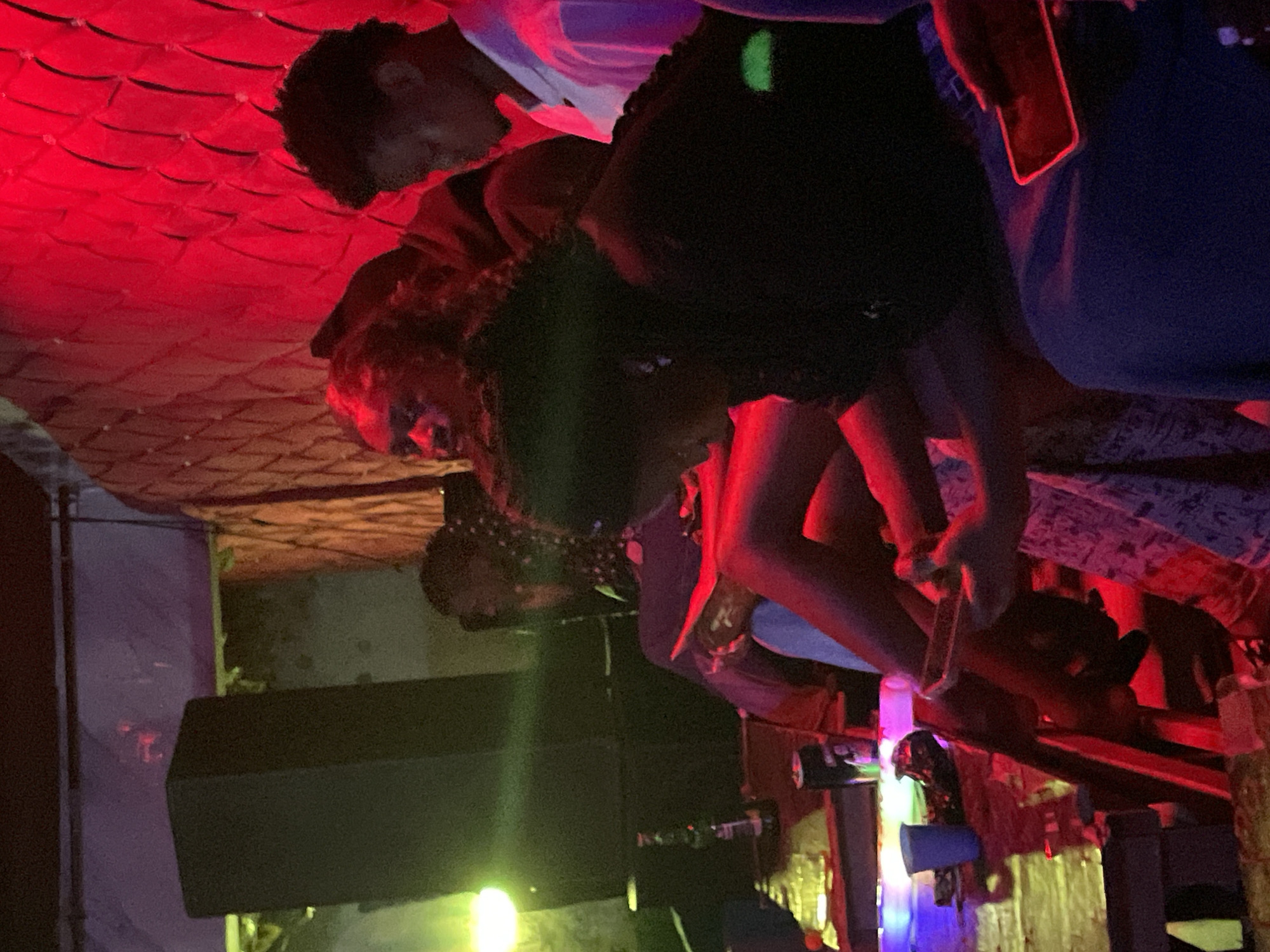 The image depicts a dimly lit interior, likely a bar or club, with predominantly red lighting. In the foreground, several individuals are seated at a table. A person on the right is holding a phone with a red case. To their left, another individual is also engaged with their phone. In the middle ground, a person with dark curly hair is partially visible, looking down. Further to the left, another person is seated, with a drink can and a bottle on the table in front of them. A large speaker is visible in the background on the left. The wall behind the individuals is textured and illuminated with red light. There is no discernible indication that the image was taken in Durimi, Nigeria.