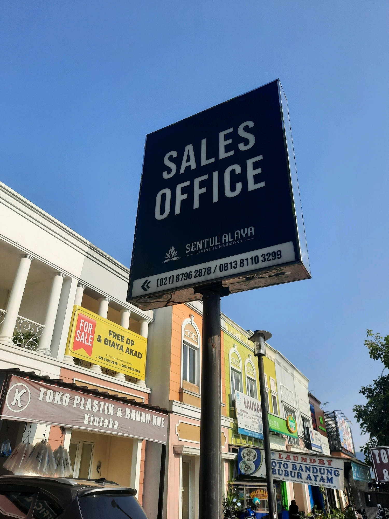 The image depicts a street scene in Babakan Madang, Indonesia, under a clear blue sky. Dominating the foreground is a tall, dark blue sign with white lettering that reads "SALES OFFICE" at the top, followed by "SENTUL ALAYA LIVING IN HARMONY" and a phone number. Below this, a yellow sign advertises "FOR SALE FREE DP & BIAYA AKAD". Further down, a maroon banner reads "TOKO PLASTIK & BAHAN KUE Kintaka". To the right, buildings with colorful facades are visible, featuring signs for "LAUNDRY" and what appears to be a food establishment selling "BUBUR AYAM". A streetlight stands between the buildings. In the lower part of the image, the rear of a dark-colored car is visible. The overall environment suggests a commercial area with shops and offices.