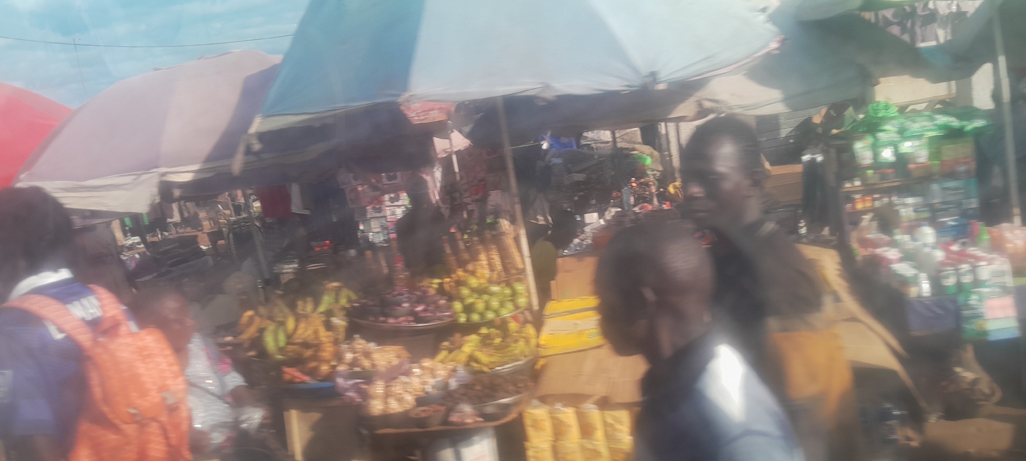 This is a daytime, medium-shot, eye-level photograph of an outdoor market scene in Nyanya, Nigeria. The market appears to be situated under a series of awnings or tarps, providing shade. In the foreground, a person wearing an orange backpack is visible from the side, their body angled to the right. To the right of this individual, another person is partially visible, facing left.
The central area of the market displays a variety of goods. There are stalls with fruits and vegetables, including bananas, green round fruits, and possibly onions or other produce. Some items are arranged in bowls, while others are presented on shelves or in containers. Further into the background, there are shelves stocked with various packaged goods, including what appear to be cleaning products or detergents, identifiable by their brightly colored packaging.
The sky is visible at the top of the image and appears to be bright and possibly overcast. The overall impression is of a bustling local marketplace with a diverse array of products being offered for sale.