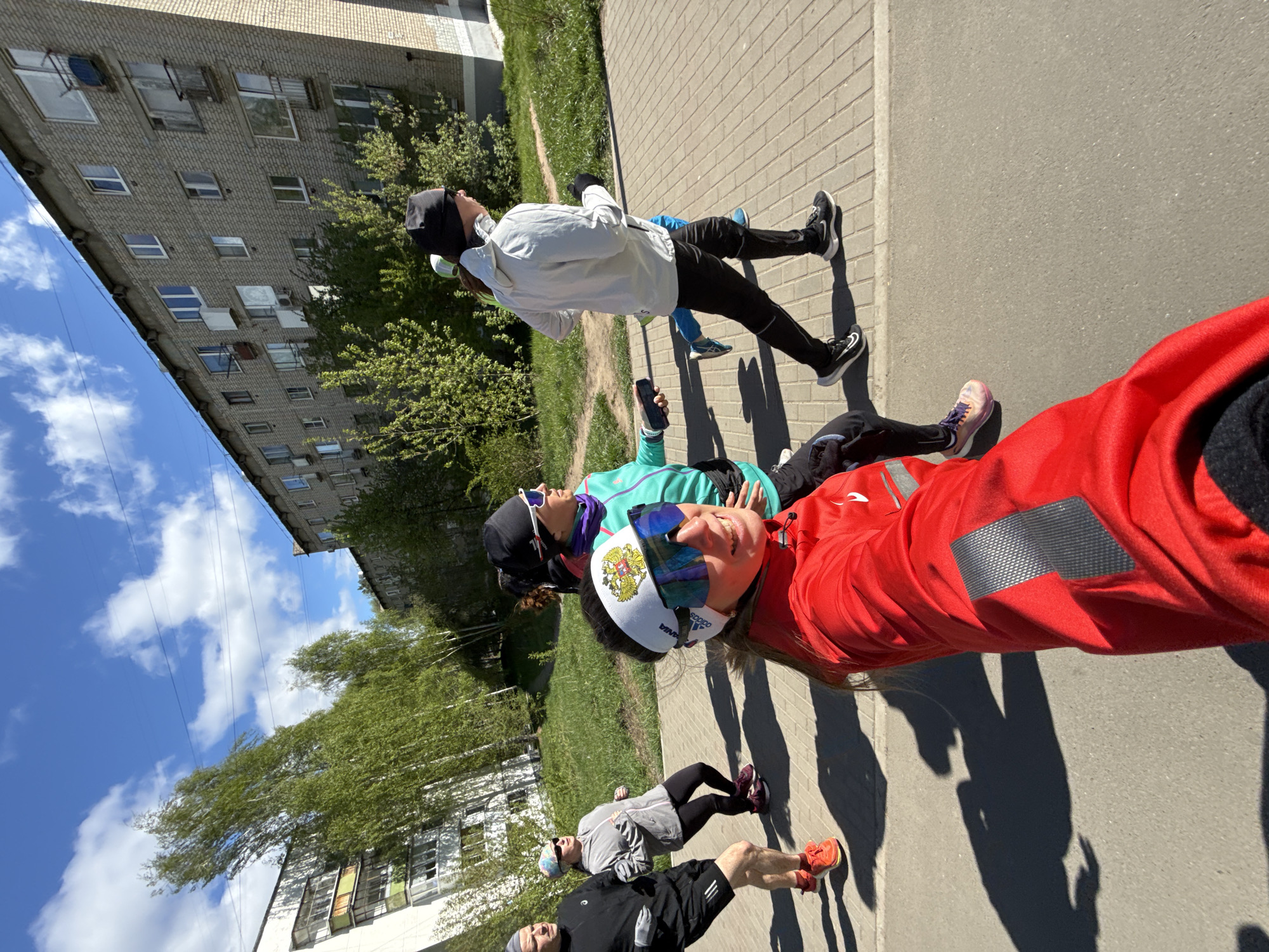 On a sunny day in Chaikovsky, Russia, a selfie captures four individuals standing on a sidewalk adjacent to a grassy area and a multi-story brick building. The person in the foreground wears a red jacket, a white headband with the Russian flag, and sunglasses. To their left, a person in a teal jacket and sunglasses holds a black smartphone. Behind them stands a person in a white jacket and a black beanie, holding a water bottle. A fourth person, wearing gray and black running attire, is visible towards the bottom left. The sidewalk runs parallel to a grassy area and a brick building with multiple windows. Power lines are visible against a blue sky with scattered clouds.