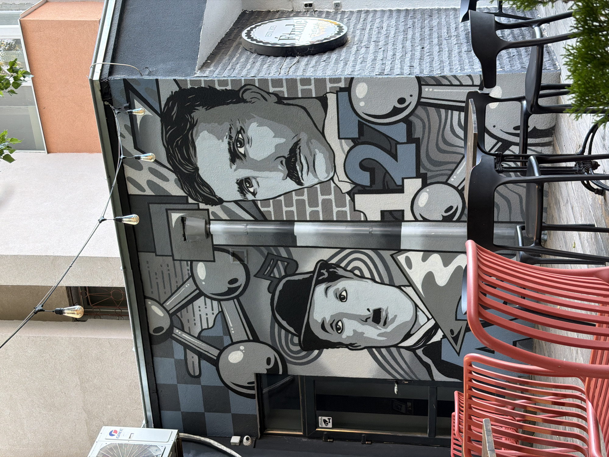 The image shows a building exterior in Blagoevgrad, Bulgaria, featuring a mural with grayscale portraits of Nikola Tesla (top) and Charlie Chaplin (bottom). Abstract geometric shapes and stylized numbers surround the portraits. A sign above the mural reads "Etude" and includes "ESTD 2003". A string of lights hangs along the edge of the building. Stacks of black and red chairs are present on the right side of the image, partially obscuring the mural. An air conditioning unit is visible at the bottom left.