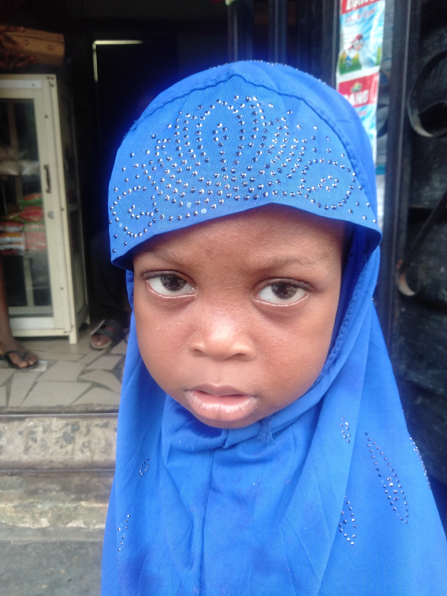A close-up portrait features a young child with brown skin, wearing a vibrant blue hijab or head covering. The hijab is adorned with numerous small, shiny silver embellishments arranged in decorative patterns across the forehead and down the sides. The child has large, dark eyes with a serious or neutral expression and slightly parted lips.
The background is somewhat blurred, suggesting a shallow depth of field. To the left of the child, a tall, narrow white cabinet with glass panels can be seen, containing various indistinct items. Further back and to the left, a person's feet in dark sandals are visible on a patterned tiled floor, indicating an entrance or doorway. To the right of the child, the background is darker, possibly an interior space or a shadowed area, with faint outlines of what appears to be a metallic gate or railing. Above the child's right shoulder, part of a colorful product wrapper or packaging is visible, bearing some text, including the partially readable word "DAN" and what might be "GANG" or "CANG". The setting appears to be an urban or semi-urban environment in Shangisha, Nigeria, likely an entrance to a shop or residence. The lighting suggests it is daytime.