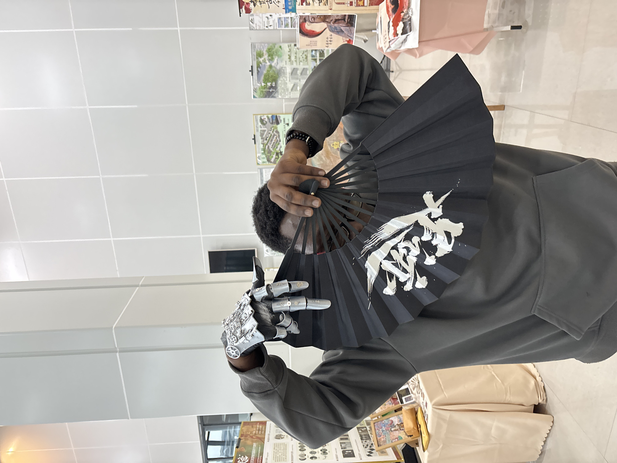 A person wearing a gray hooded sweatshirt holds a black fan with white calligraphy in front of their face, partially obscuring it. The person wears a silver, mechanical-looking glove on their left hand, which is holding the fan. The background includes a white tiled wall and floor. Several posters or pictures are visible behind the person, along with a table covered with a light-colored cloth. The location is indoors and appears to be an exhibition or event space. No evidence in the image identifies the location as Jiangjin, China.