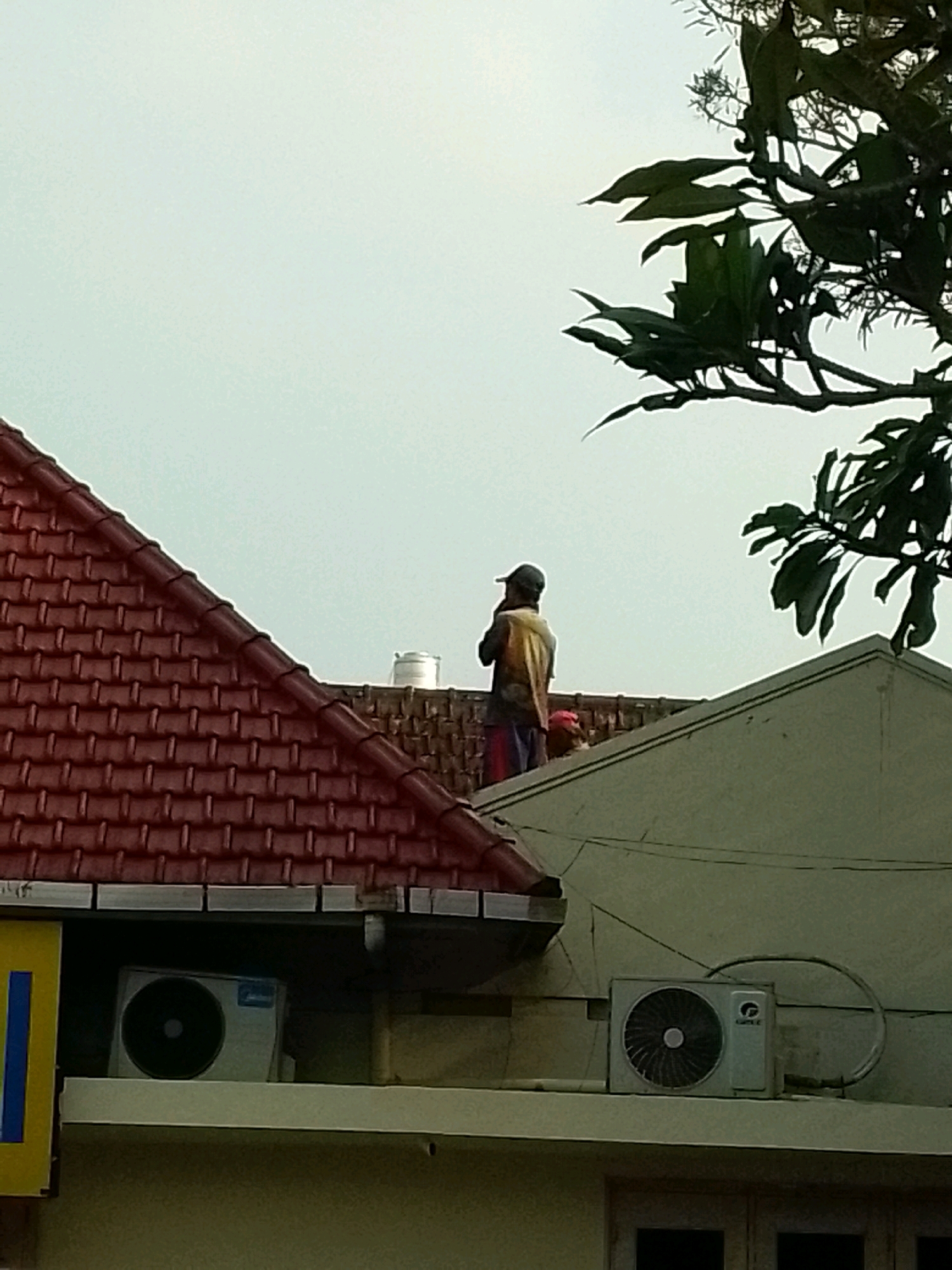 A person stands on a rooftop in Jember, Indonesia, silhouetted against an overcast sky. The person is wearing a cap, a light-colored jacket over a darker shirt, and striped trousers. They appear to be holding a phone to their ear, suggesting they are engaged in a conversation. In the foreground, a red-tiled roof slopes downwards, while to the right, a light-colored roof leads to a section of a building. Two outdoor air conditioning units are visible on ledges below the roofs. Green foliage from a tree extends into the upper right corner of the frame. The scene suggests a typical urban environment with residential and commercial buildings.