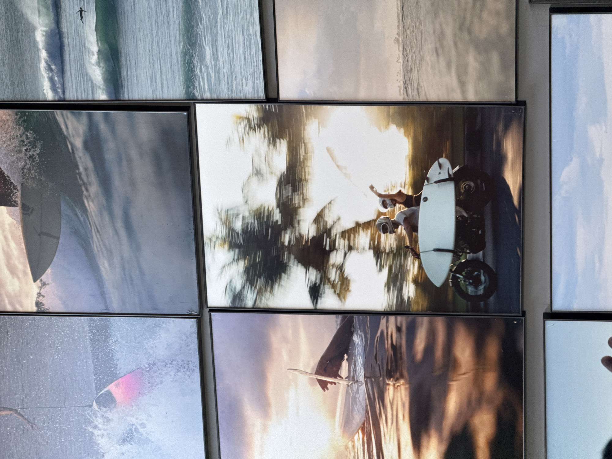 A collection of framed photographs hanging on a wall, likely in Kuta Selatan, Indonesia, given the subject matter. The photos primarily depict scenes related to surfing and coastal landscapes. Visible subjects include surfers riding waves, a photograph of a person on a motorcycle with a surfboard attached, possibly palm trees, and ocean waves. Actions captured involve surfing, and the environment suggests a beach or coastal area. The objects include surfboards, motorcycles, and framed photographs.