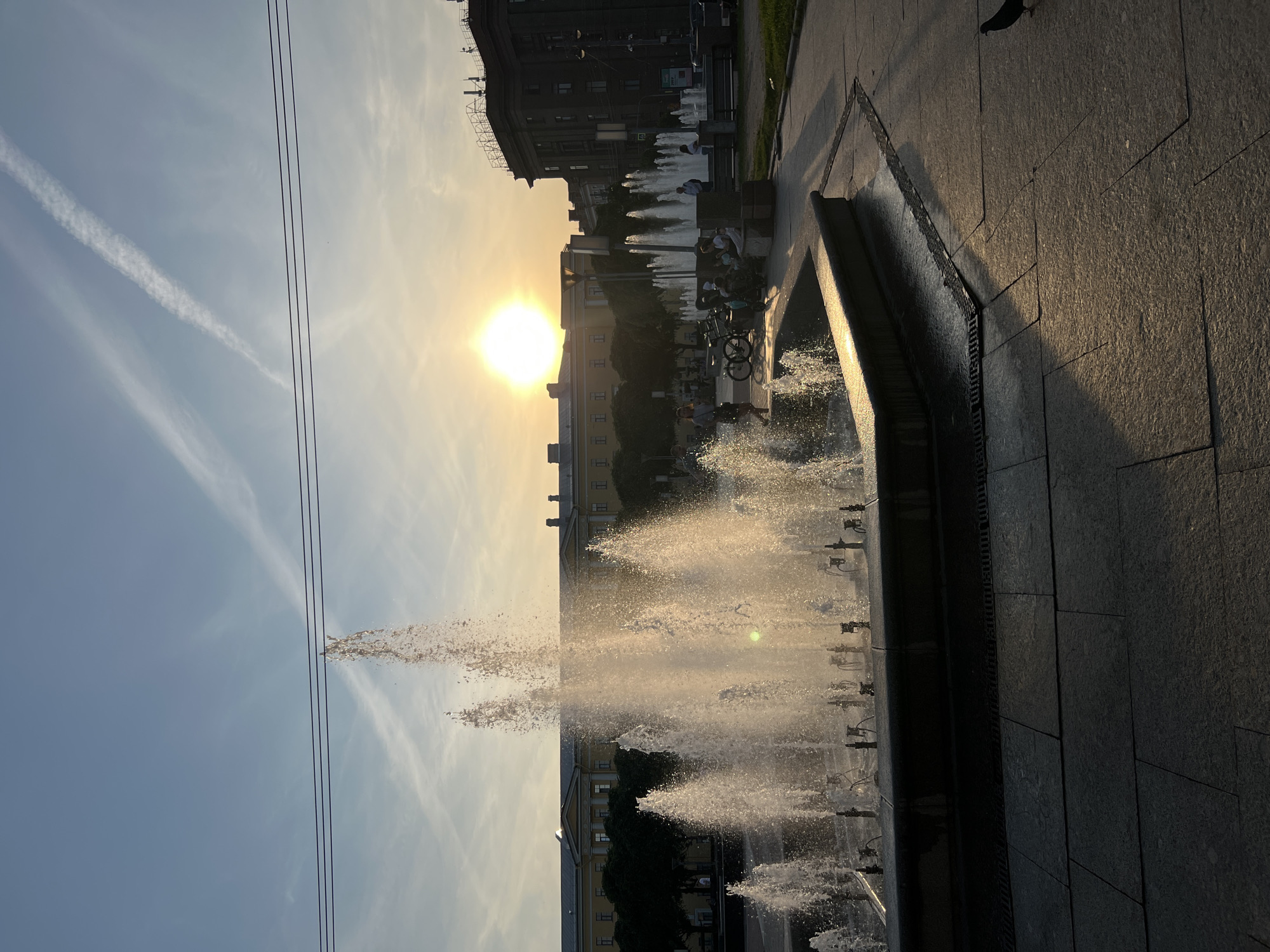 The image depicts a public square in Saint Petersburg, Russia, during a sunset. A large fountain is in the foreground, with water spraying upwards and catching the warm light of the setting sun. Several people are visible, some sitting on a bench near the fountain, and others walking in the background. A bicycle is parked nearby. The architecture of the buildings surrounding the square suggests a historical urban setting. The sky is clear with some wispy clouds, and power lines are visible across the upper portion of the frame. The ground is paved with large stone tiles, reflecting the golden light of the sunset.